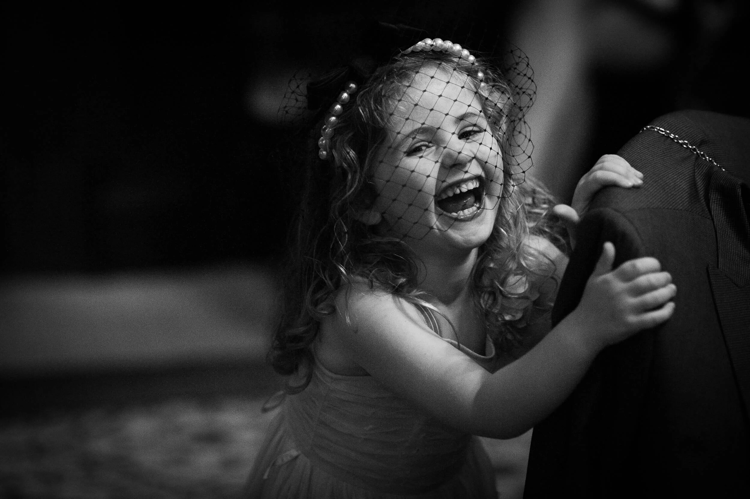 A young girl in a dress and a veil, joyfully smiling and laughing while holding onto a person dressed in dark clothing.