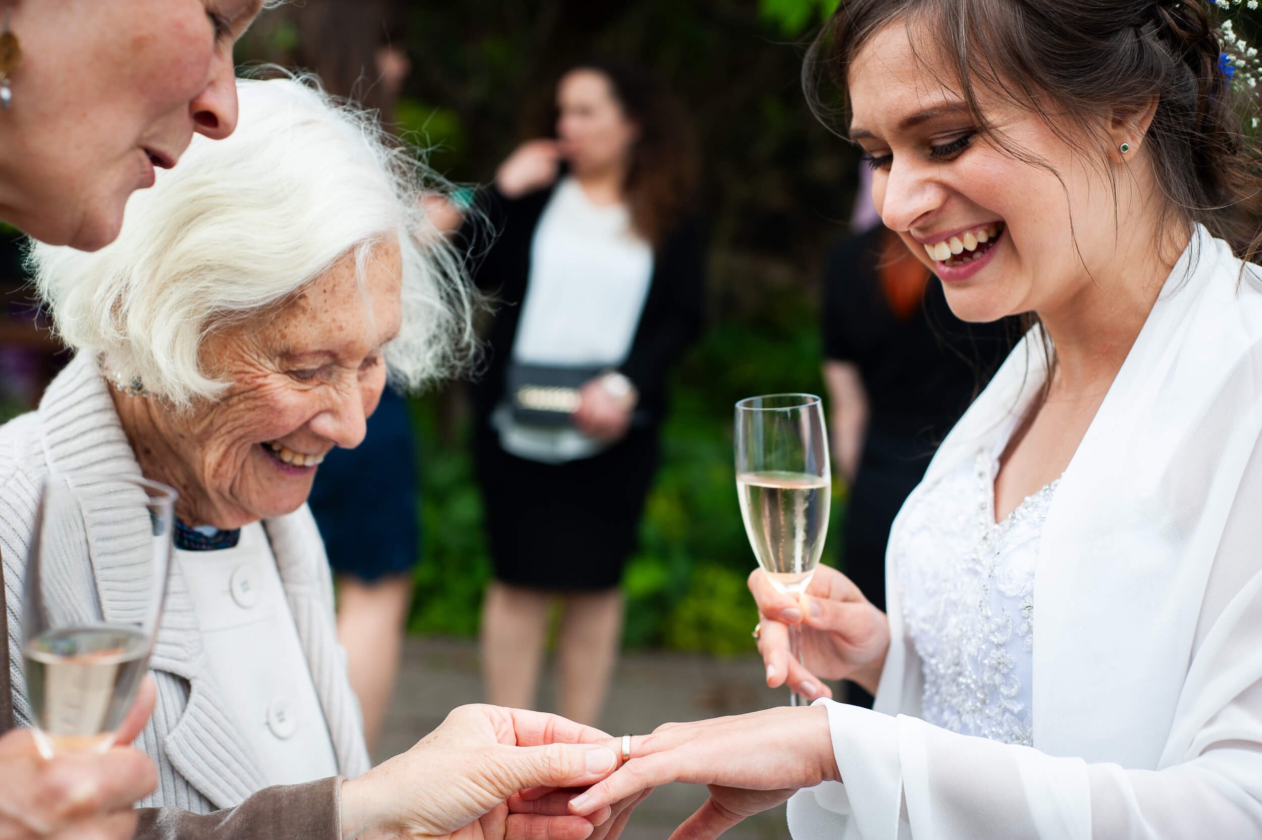 A woman gives a ring to an older woman during a celebration, with friends holding glasses of champagne in a garden setting.