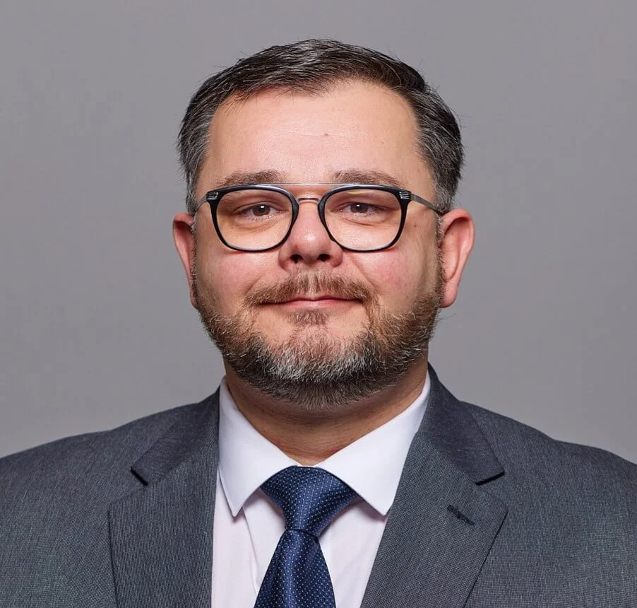 Headshot of a man with glasses, a beard, and mustache, wearing a suit and tie against a gray background.