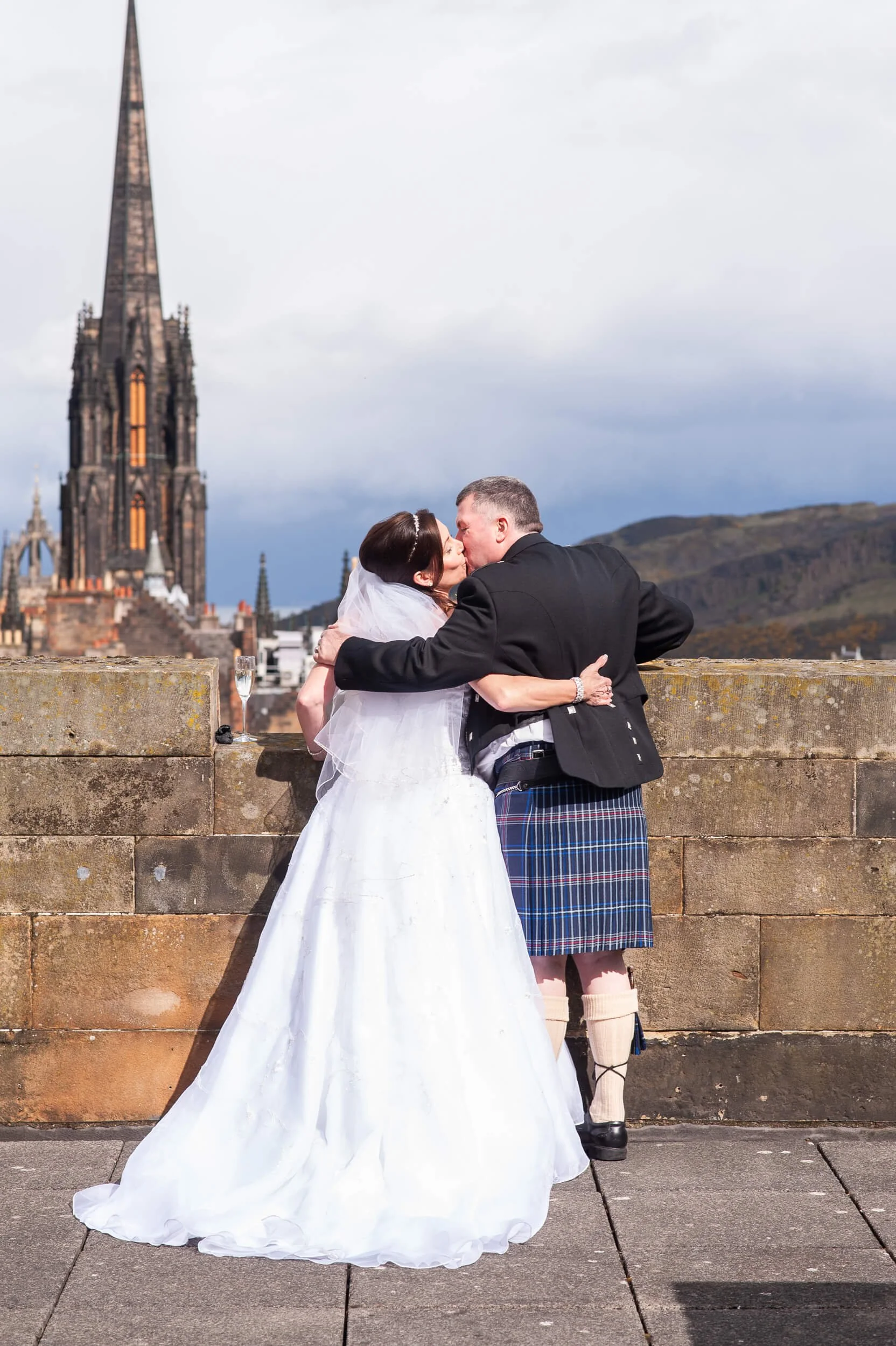 Bride and groom kissing on Edinburgh Castle ramparts with the Hub and Arthur's Seat in the background — wedding photography Scotland