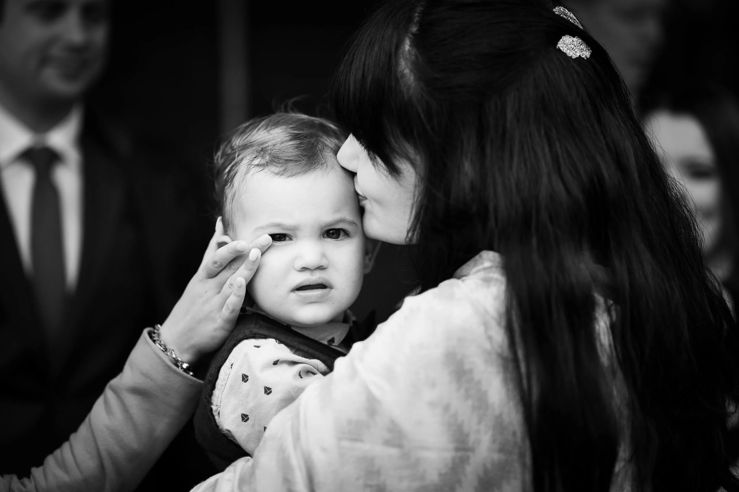 A woman holding a young boy, who has a concerned or confused expression, close to her face, with another person partially visible in the background.
