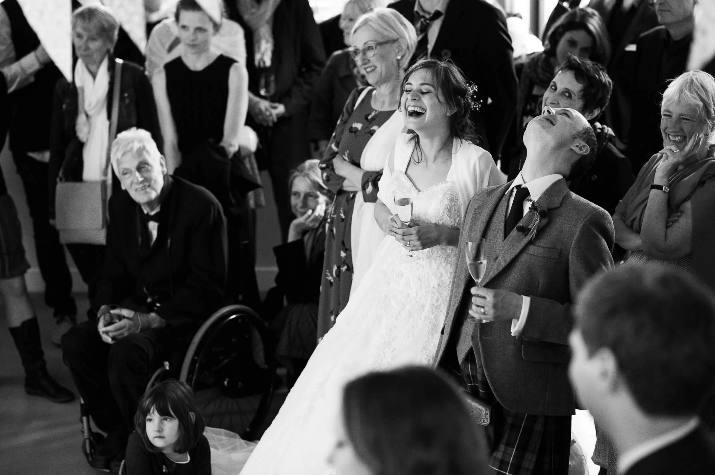 A wedding reception with guests laughing and smiling, including a bride in a white dress holding a glass, and a man in a kilt.