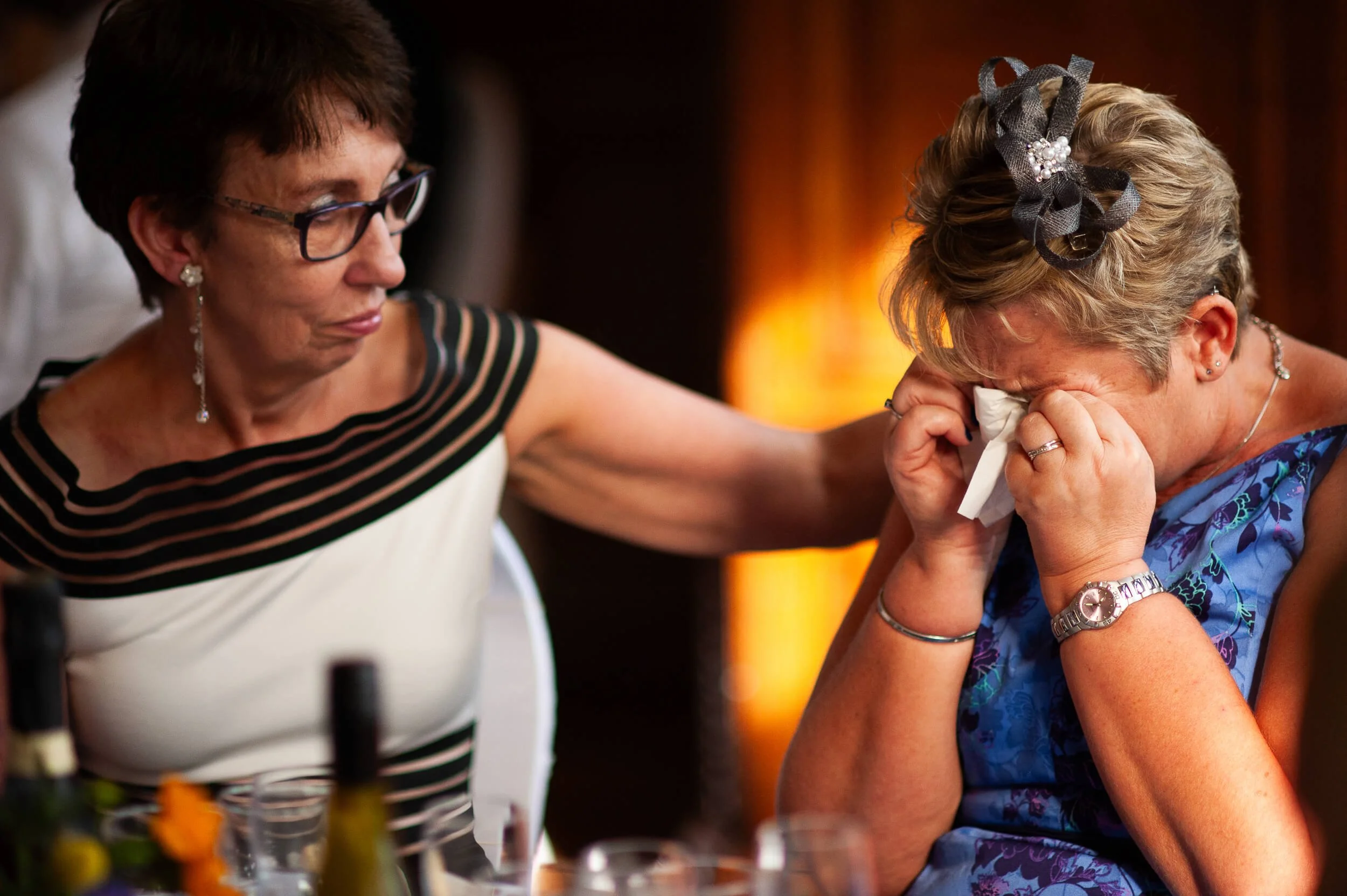 A woman in glasses and a striped dress is comforting another woman who is crying and wiping tears with a tissue, during a gathering or celebration.