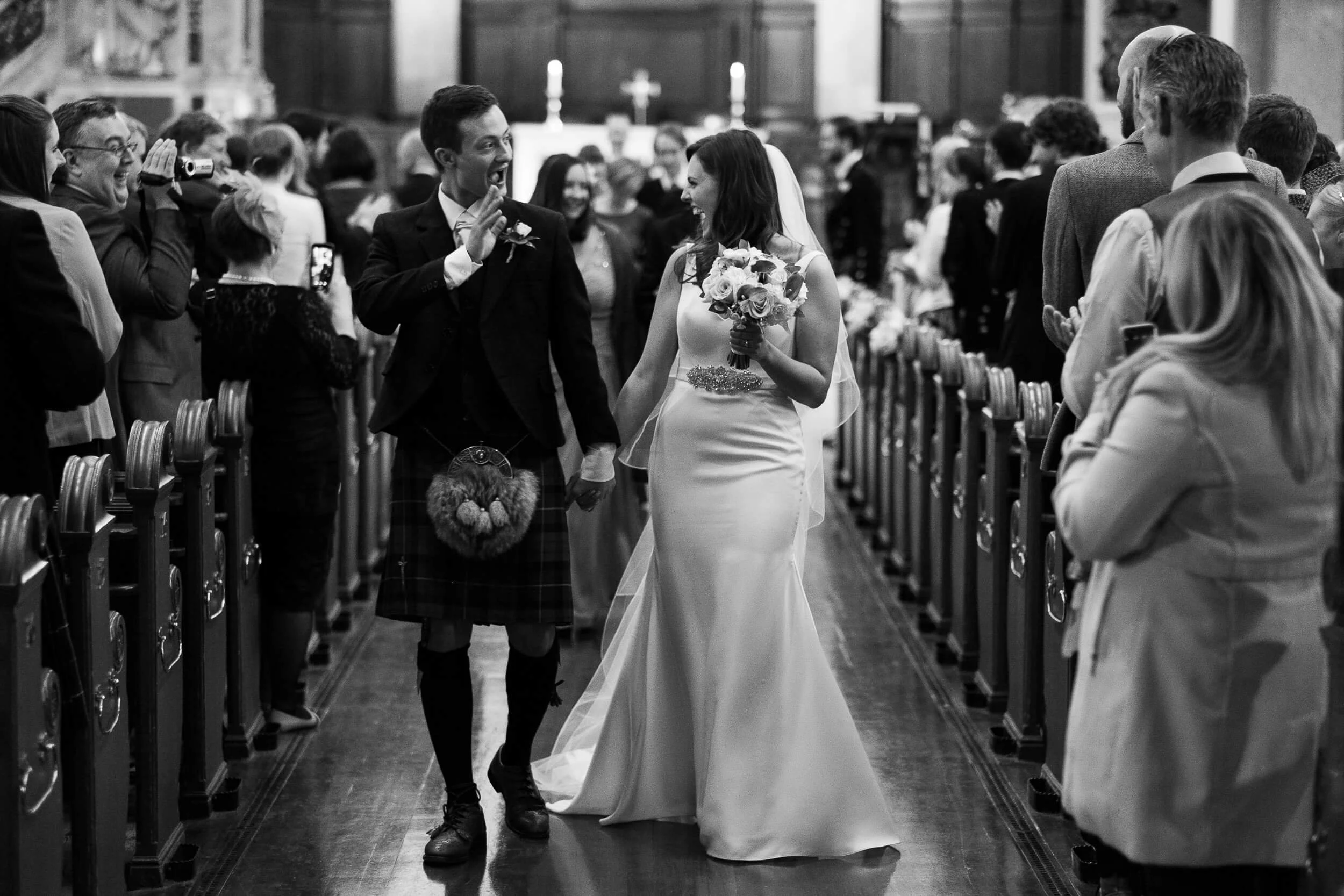 A bride and groom walking down the aisle in a church, holding hands, with the bride holding a bouquet. Guests on both sides are smiling, clapping, and taking photos of the couple.