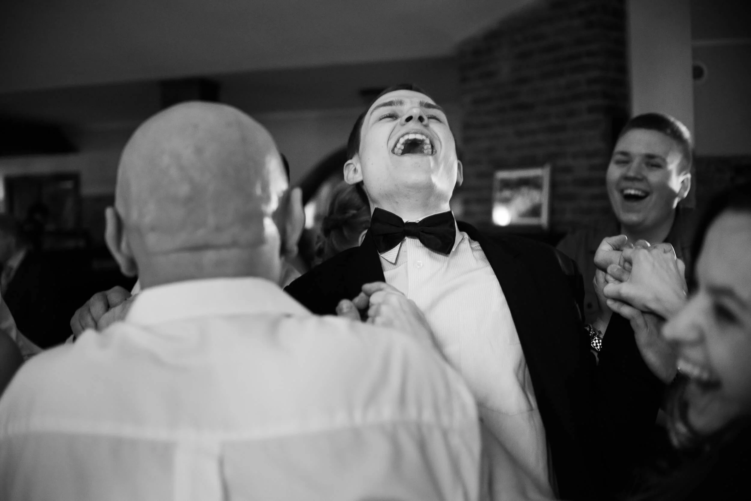 People dancing together, smiling and laughing, at a celebration or party, with a focus on a man in a tuxedo and bow tie.