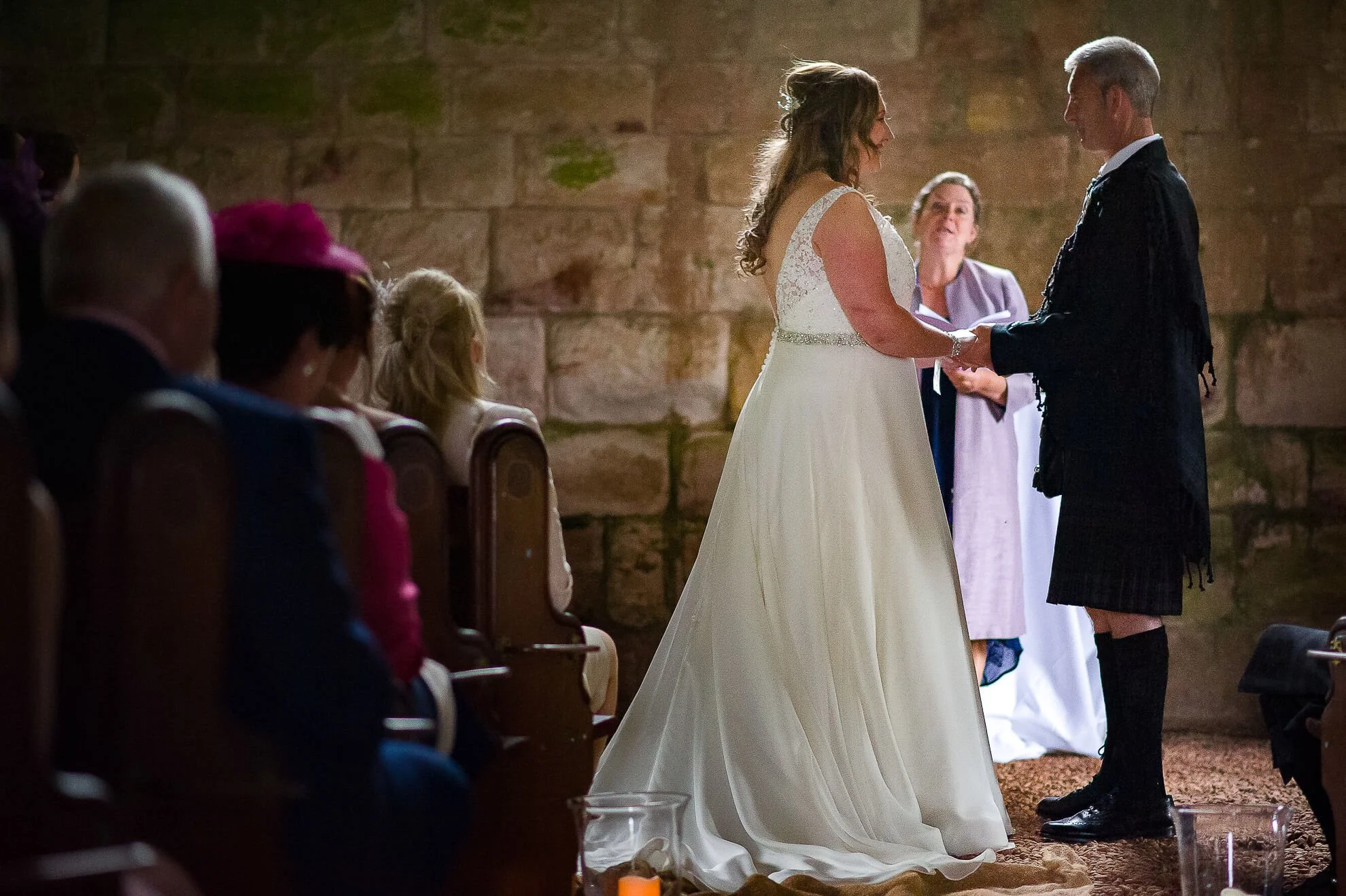 A wedding ceremony with a bride and groom holding hands, exchanging vows in a rustic setting with stone walls. Guests are seated on wooden chairs, watching the couple.
