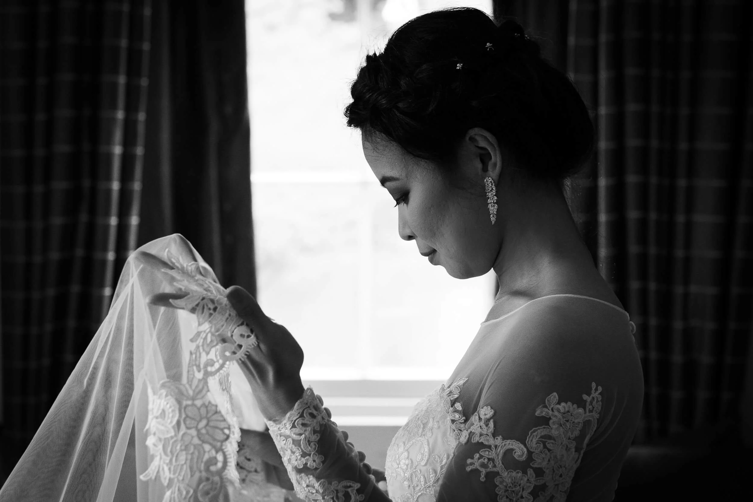 A bride in a lace wedding dress holding a veil in front of a window with dark curtains.