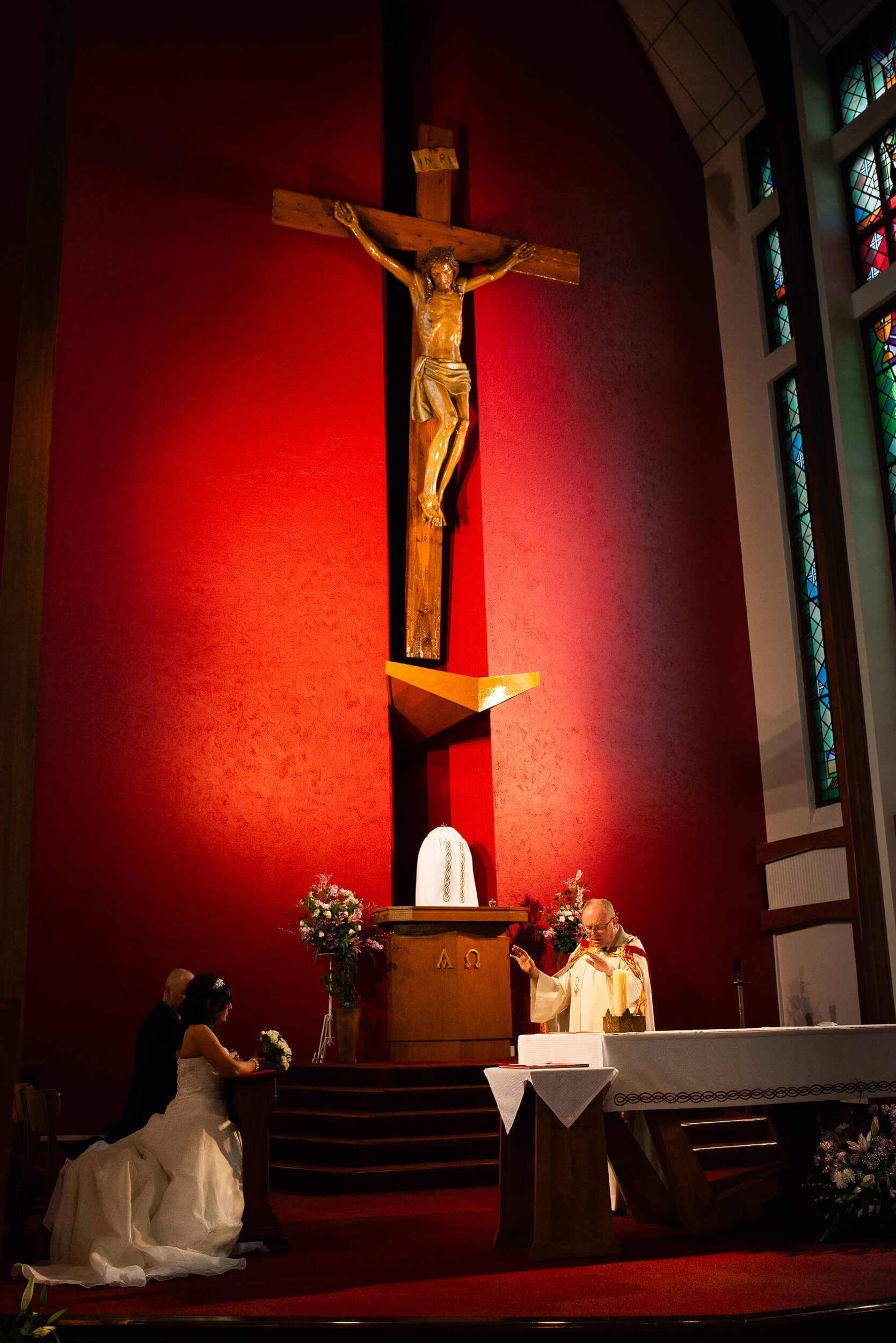 A priest conducting a wedding ceremony with a bride and groom kneeling at the altar inside a church, behind a large crucifix of Jesus on the cross, with stained glass windows visible on the side.