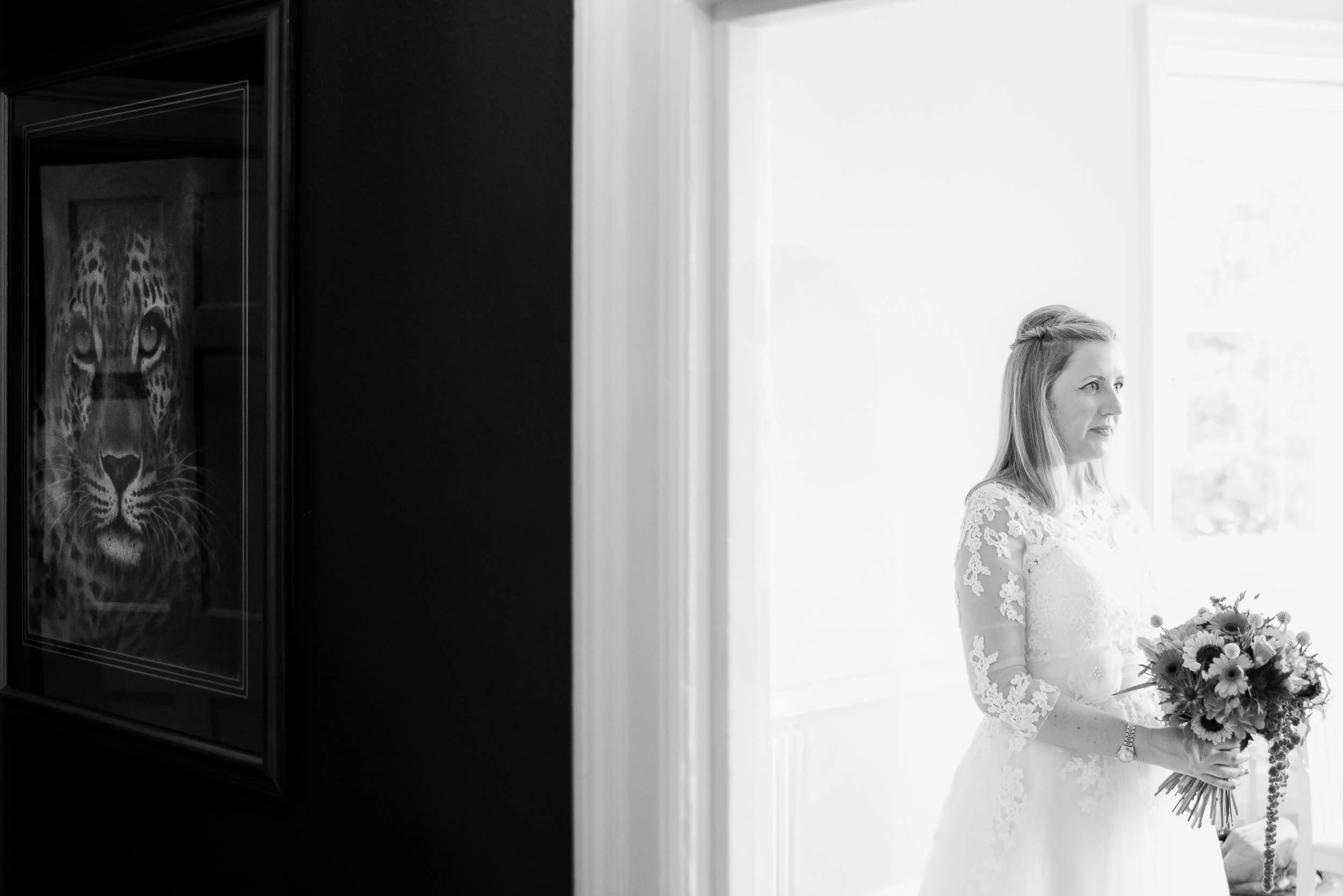 Black and white photo of a woman in a wedding dress holding a bouquet, standing near a window with light coming through. There is a framed picture of a jaguar on the dark wall beside her.
