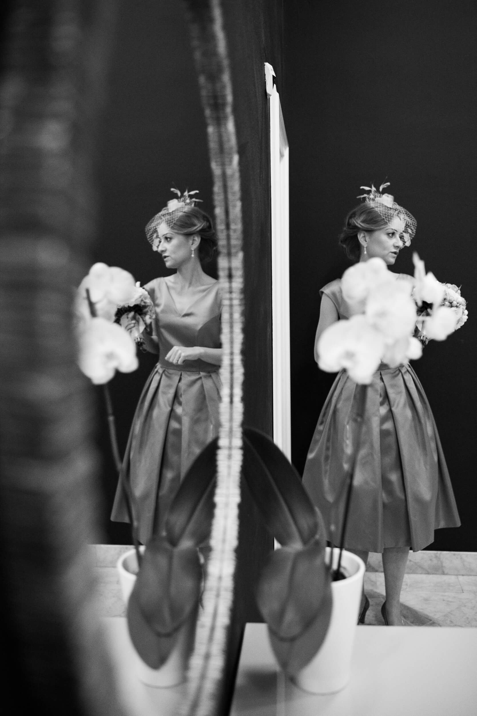 A woman in a vintage-style dress and a fascinator hat looking at her reflection in a mirror, holding a bouquet of flowers.