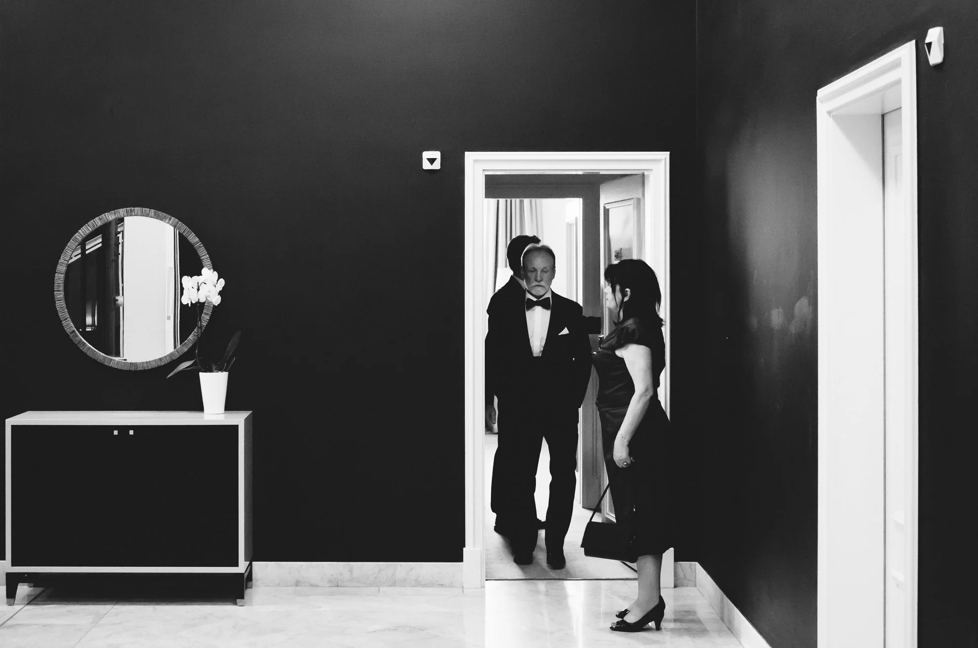 A black and white photograph of a formal setting, showing a woman in a black dress and high heels holding a handbag, standing by a dark wall and looking toward a doorway where a man in a tuxedo and a woman are entering.
