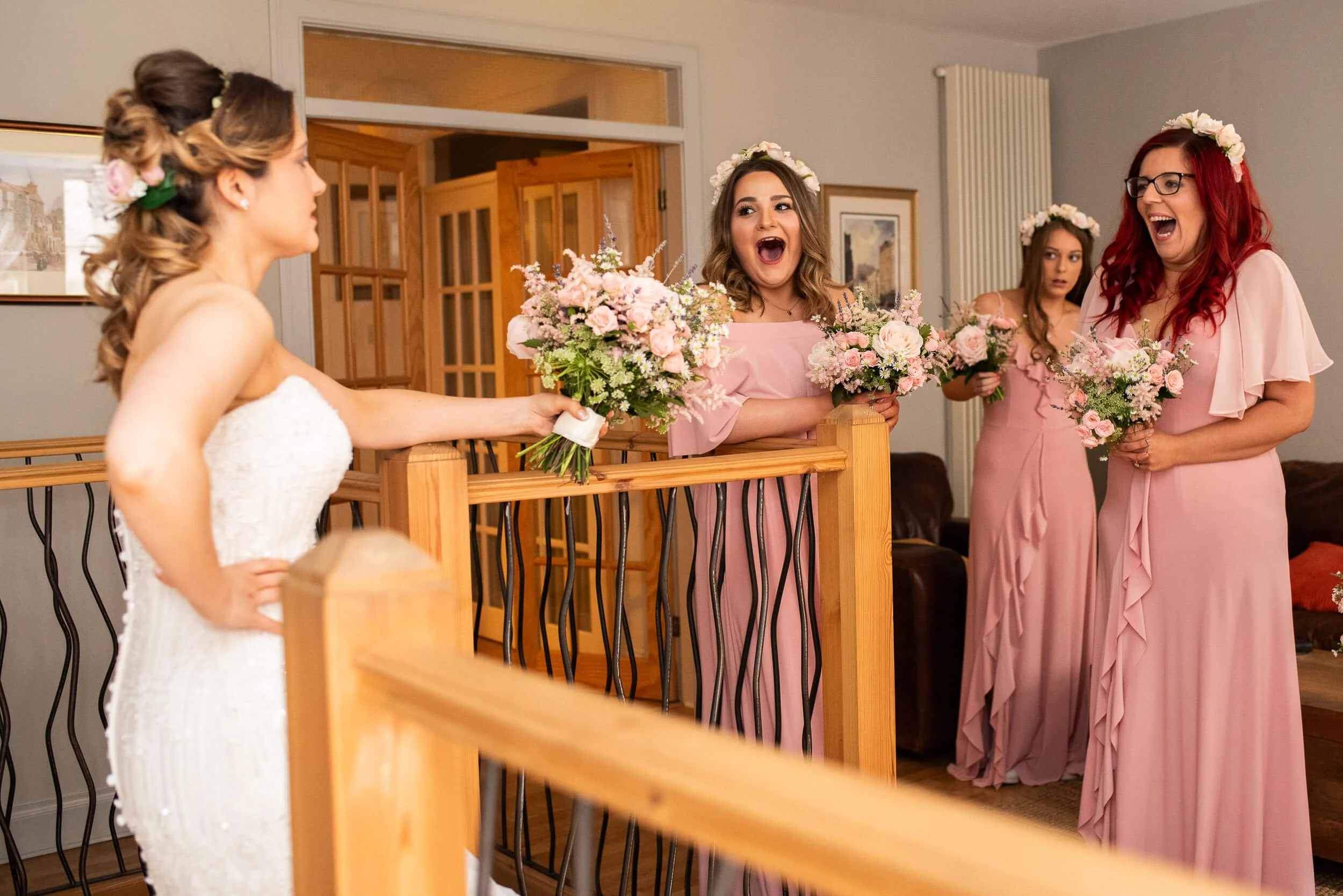 Bride in a white dress happily receiving flowers from her bridesmaids, who are dressed in pink gowns and holding bouquets at a wedding celebration.