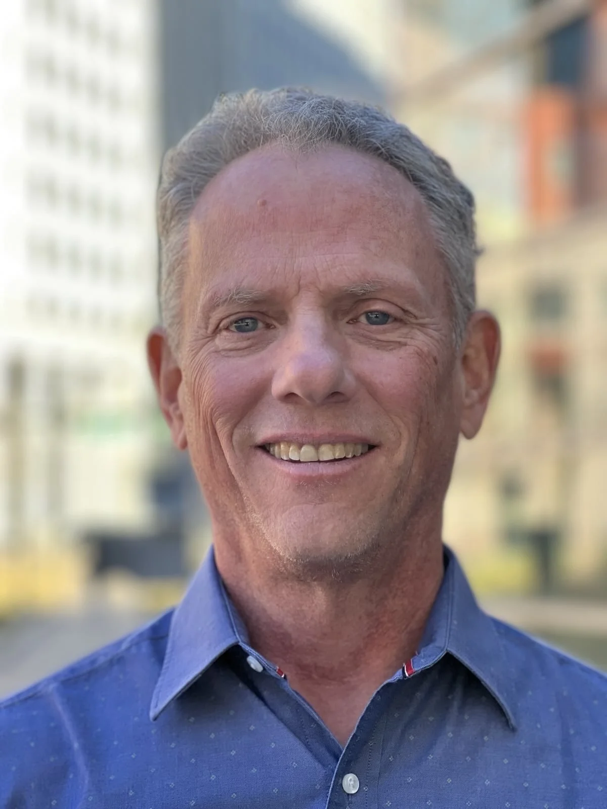 A middle-aged man with short gray hair and blue eyes, smiling and wearing a blue button-up shirt, with a blurred cityscape background.