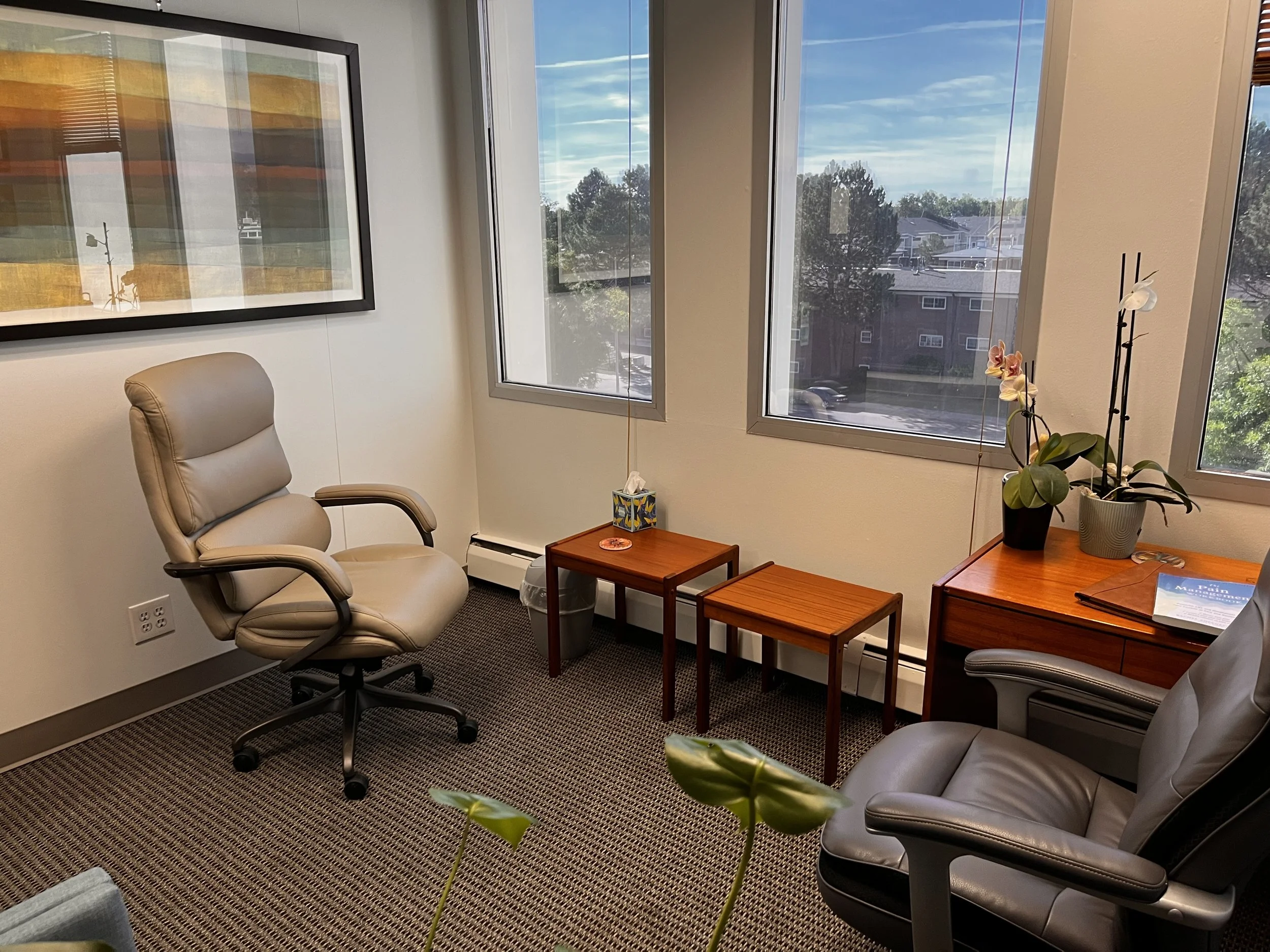 Office room with beige chairs, small wooden tables, potted orchid plant, large windows with a view of trees and buildings outside.In Person Therapy room for Good Enough Counseling, Denver