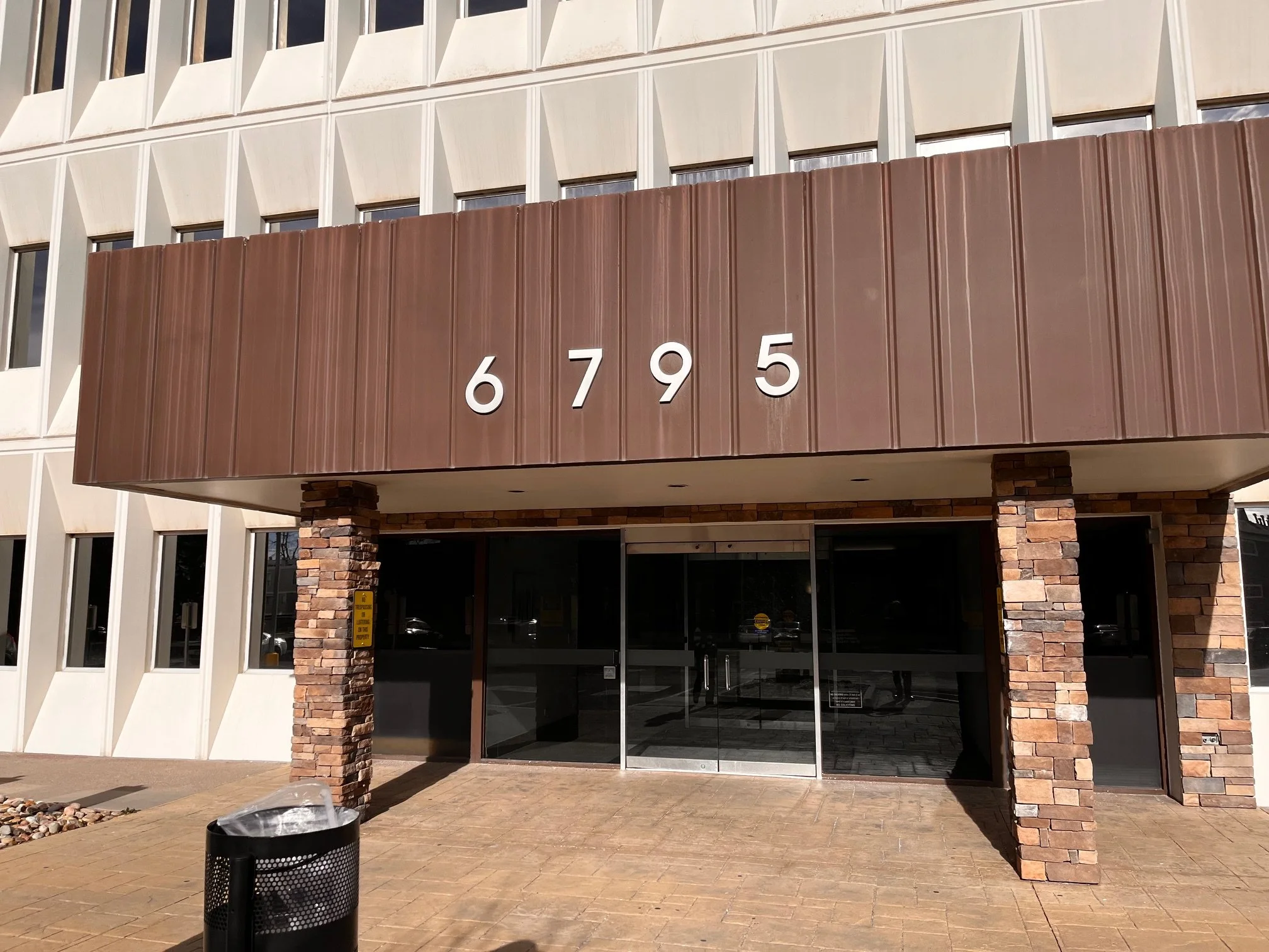 Building entrance with the number 6795 displayed on a brown metal awning, brick columns, and glass doors, with a trash can in the foreground.