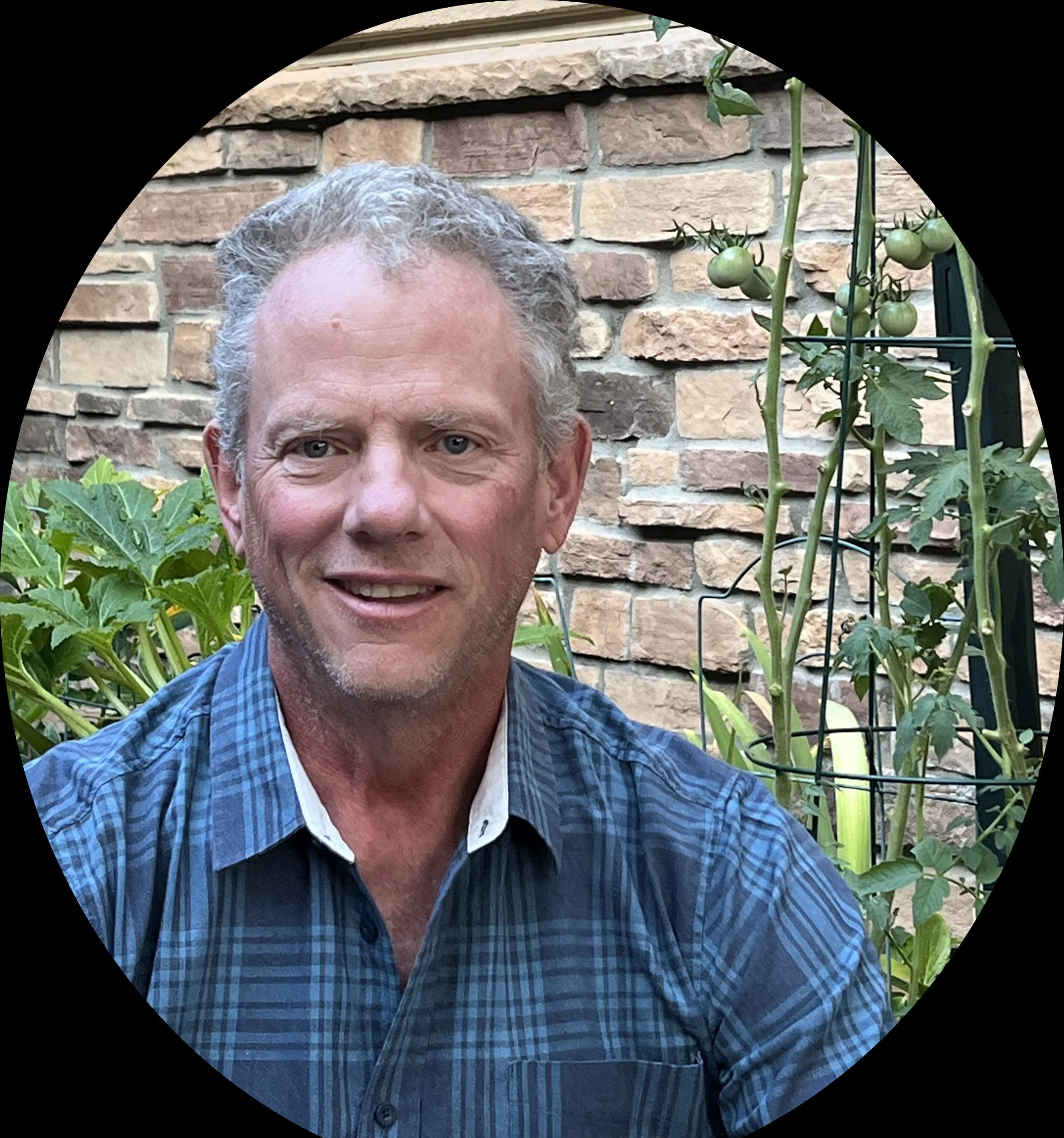Close-up of a middle-aged man with gray hair and blue eyes, smiling, wearing a blue plaid shirt, standing outdoors by a brick wall with green tomato plants.