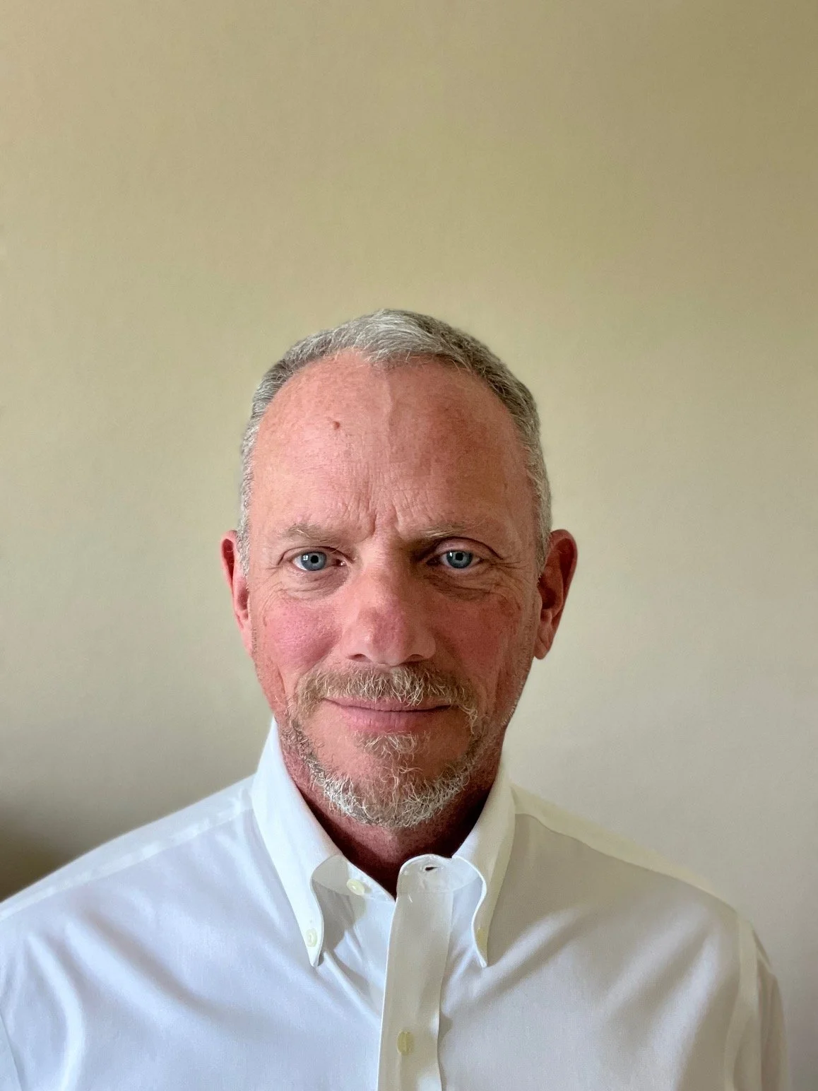 Close-up portrait of a middle-aged man with blue eyes, short gray hair, a beard, wearing a white collared shirt, with a neutral beige background. Brian Whitney, Therapist