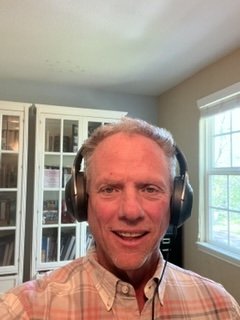 A middle-aged man with gray hair wearing large headphones, smiling, and sitting in a room with white bookshelves and a window with blinds. Brian Whitney LCSW