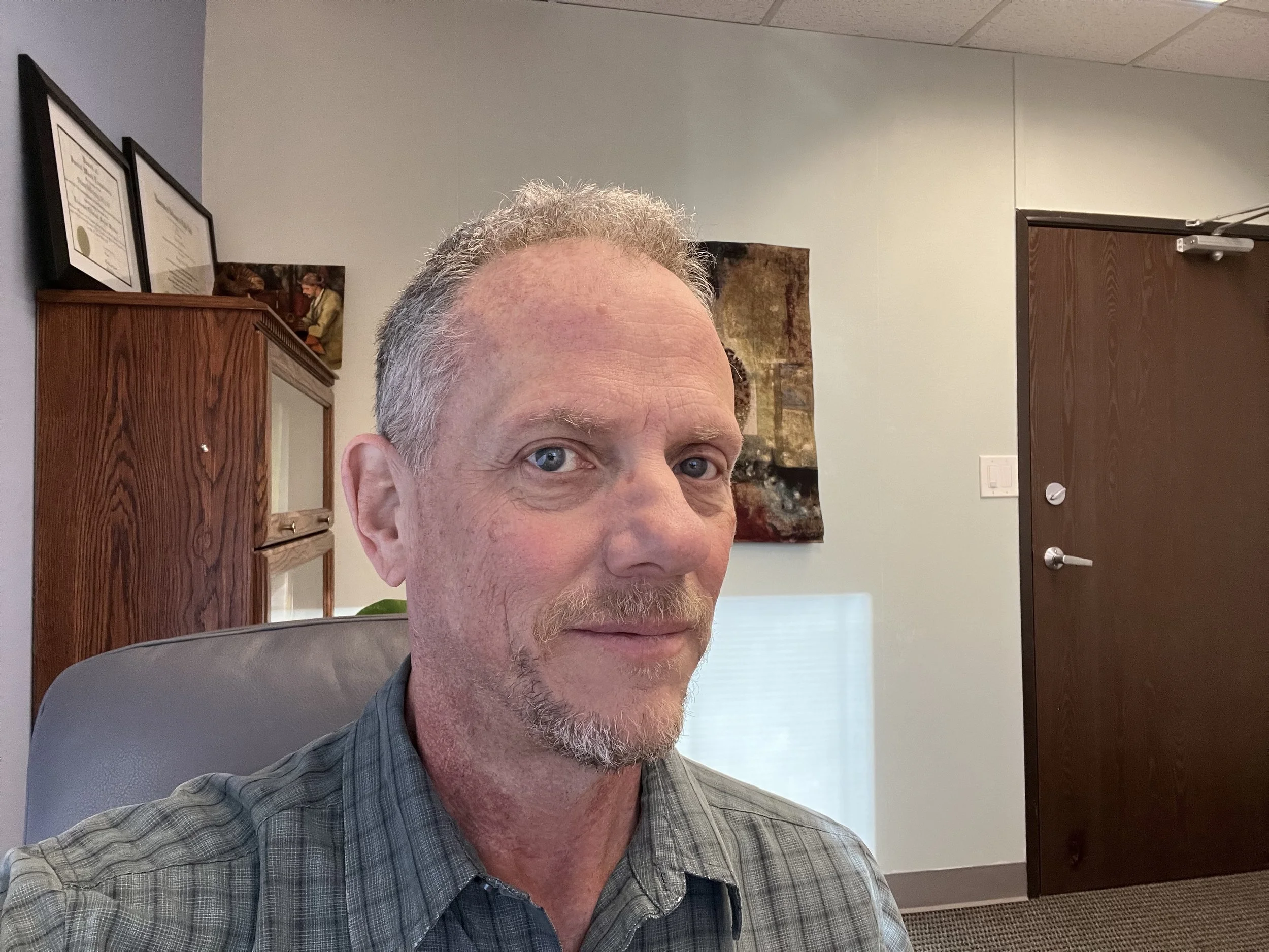 Close-up of middle-aged man with gray hair and blue eyes, wearing a gray plaid shirt, sitting in an office with framed documents and artwork on the wall behind him, and a brown wooden cabinet.