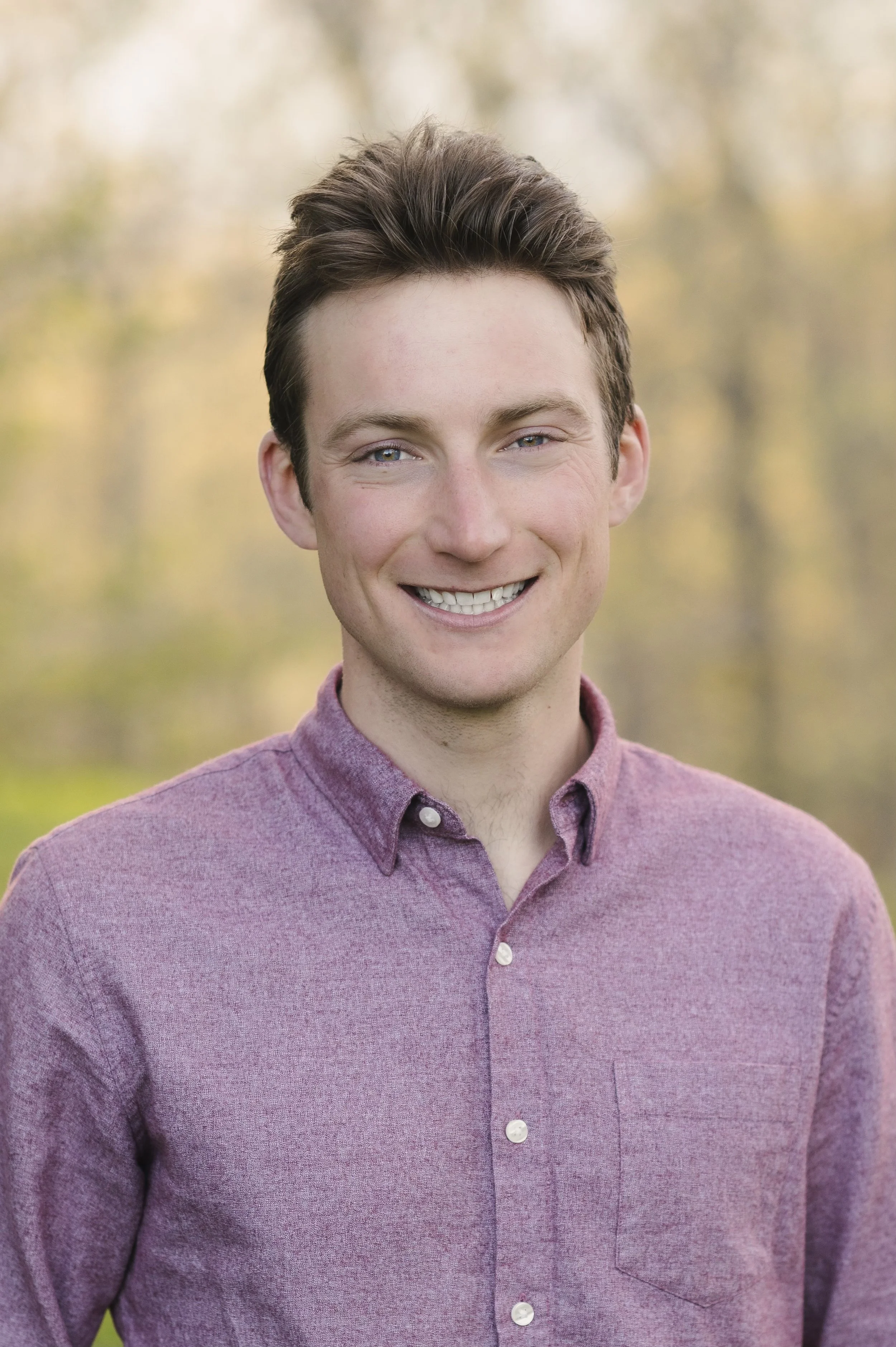 Headshot of a smiling young man with brown hair, wearing a maroon button-up shirt, outdoors with blurred trees in the background.