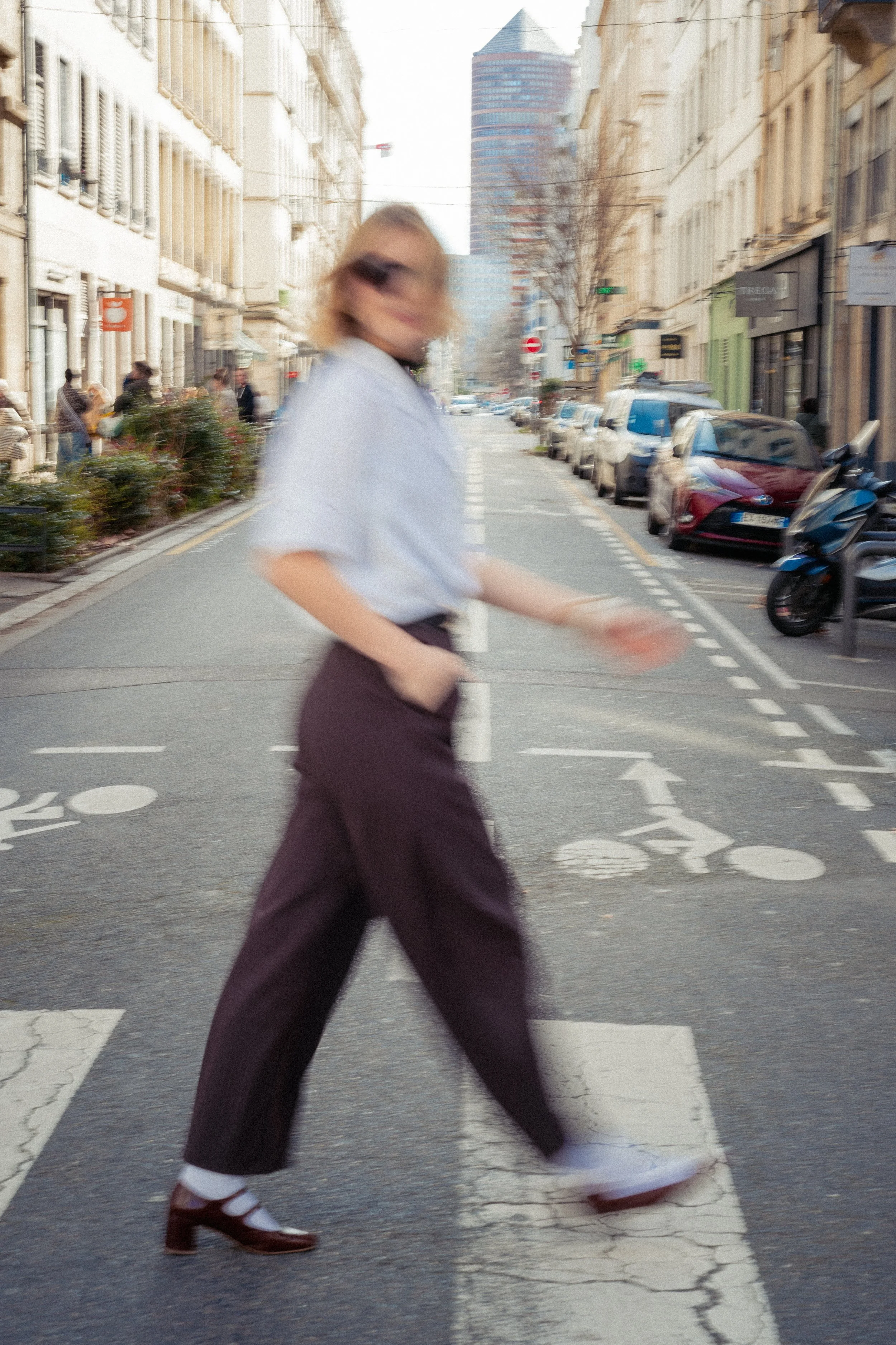 Une personne traversant un passage pour piétons dans une rue urbaine, avec des bâtiments et des voitures stationnées en arrière-plan, portant des chaussures à talons et une jupe ou pantalon sombre. Le shooting est romantique et flou.