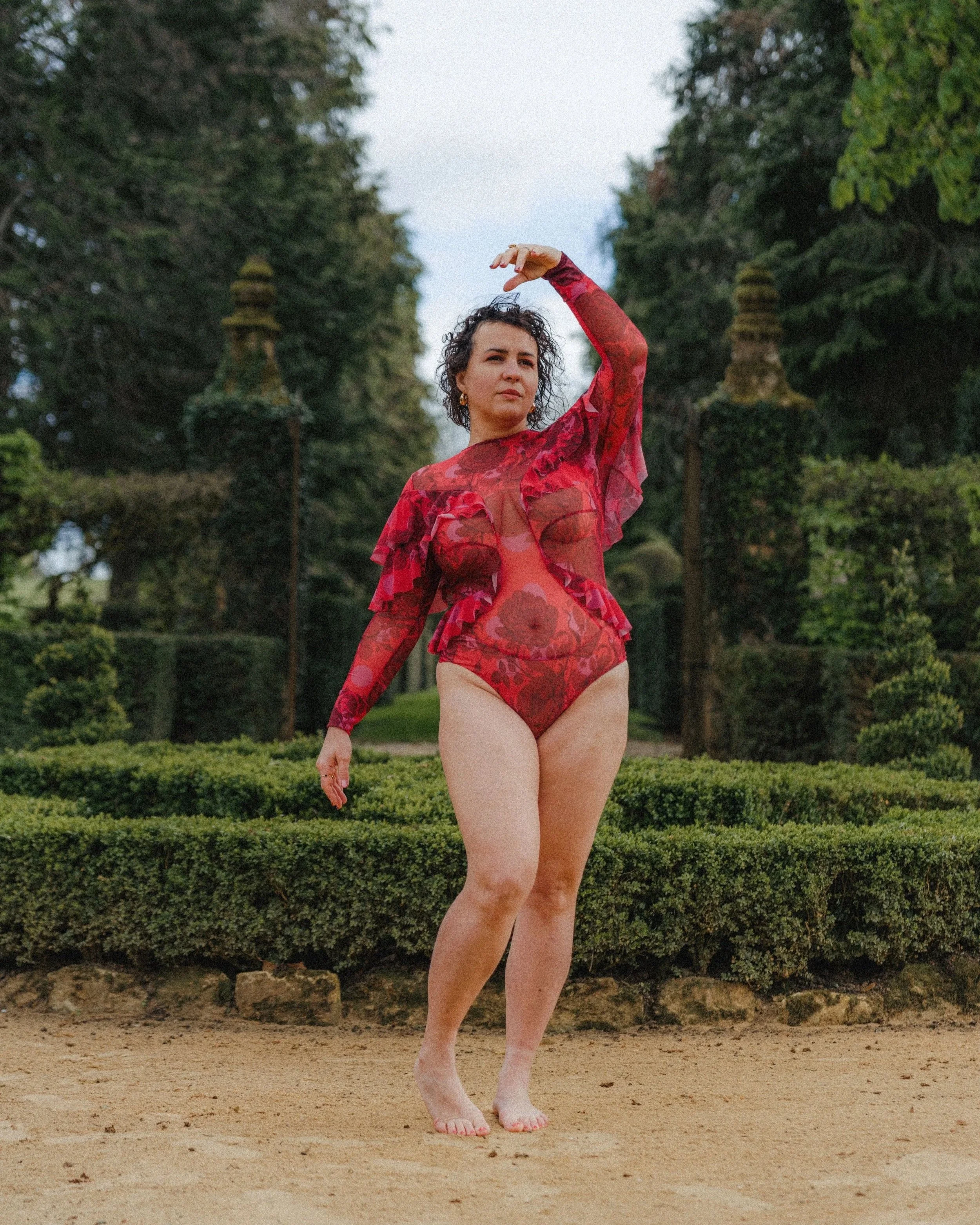 Une femme en maillot de bain à motifs rouges, debout dans un jardin avec des arbres et des buissons, en train de faire une pose artistique avec un bras levé. Elle a des cheveux bouclés et porte des boucles d'oreilles, elle regarde vers la caméra.