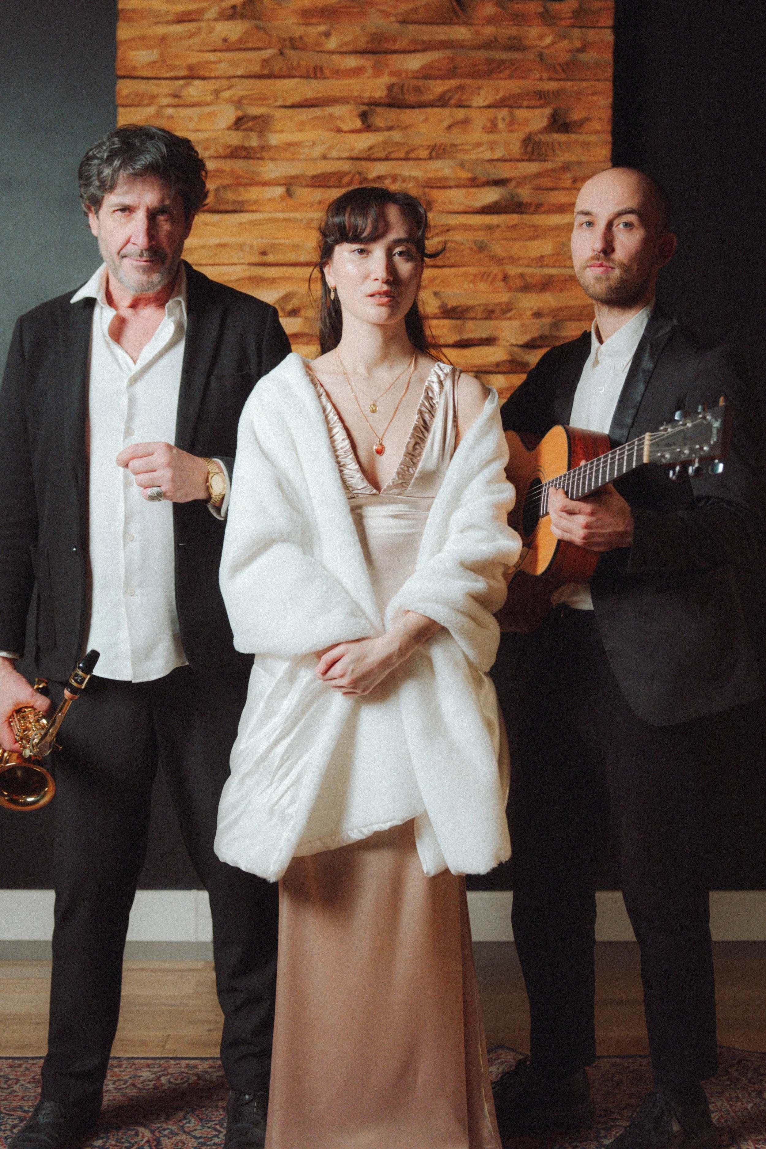 Trois musiciens dans une pièce avec un mur en bois en arrière-plan. La chanteuse se tient au centre, portant une robe blanche avec des bijoux, les hommes portent des costumes noirs, l'un avec une guitare et l'autre avec un saxophone.