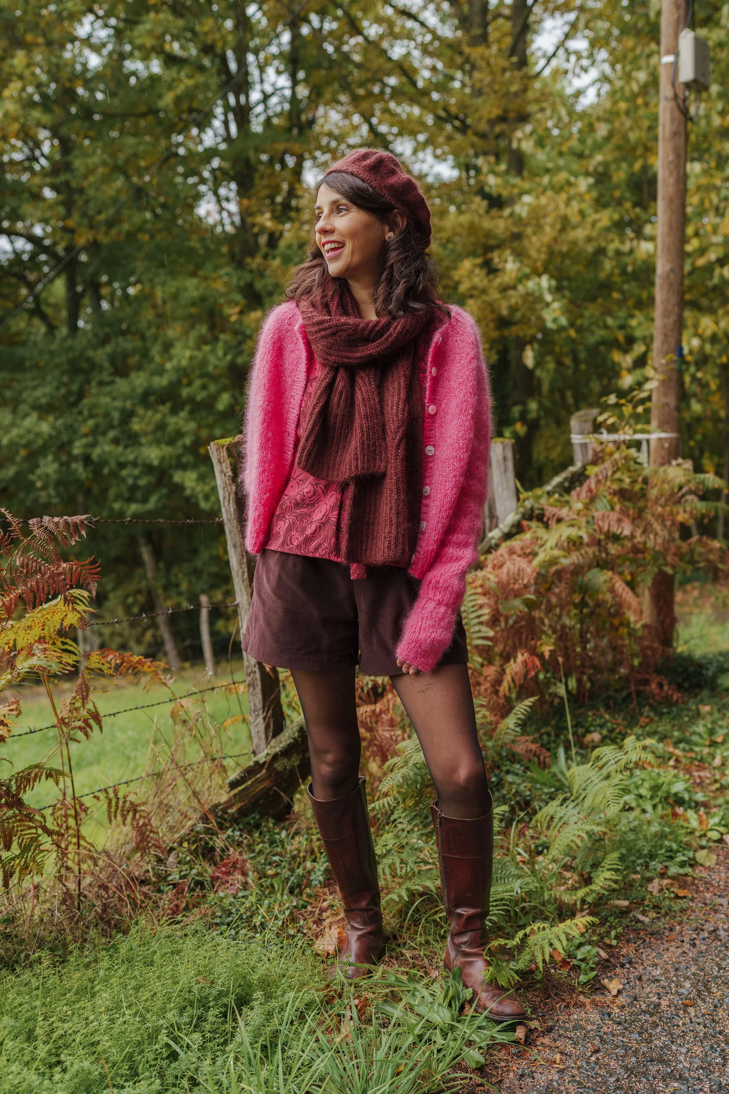 Femme souriante portant un pull rose, un bonnet marron, un foulard marron, une jupe rose avec motif, des collants noirs et des bottes en cuir marron, debout dans un environnement naturel automnal avec des arbres et des fougères.