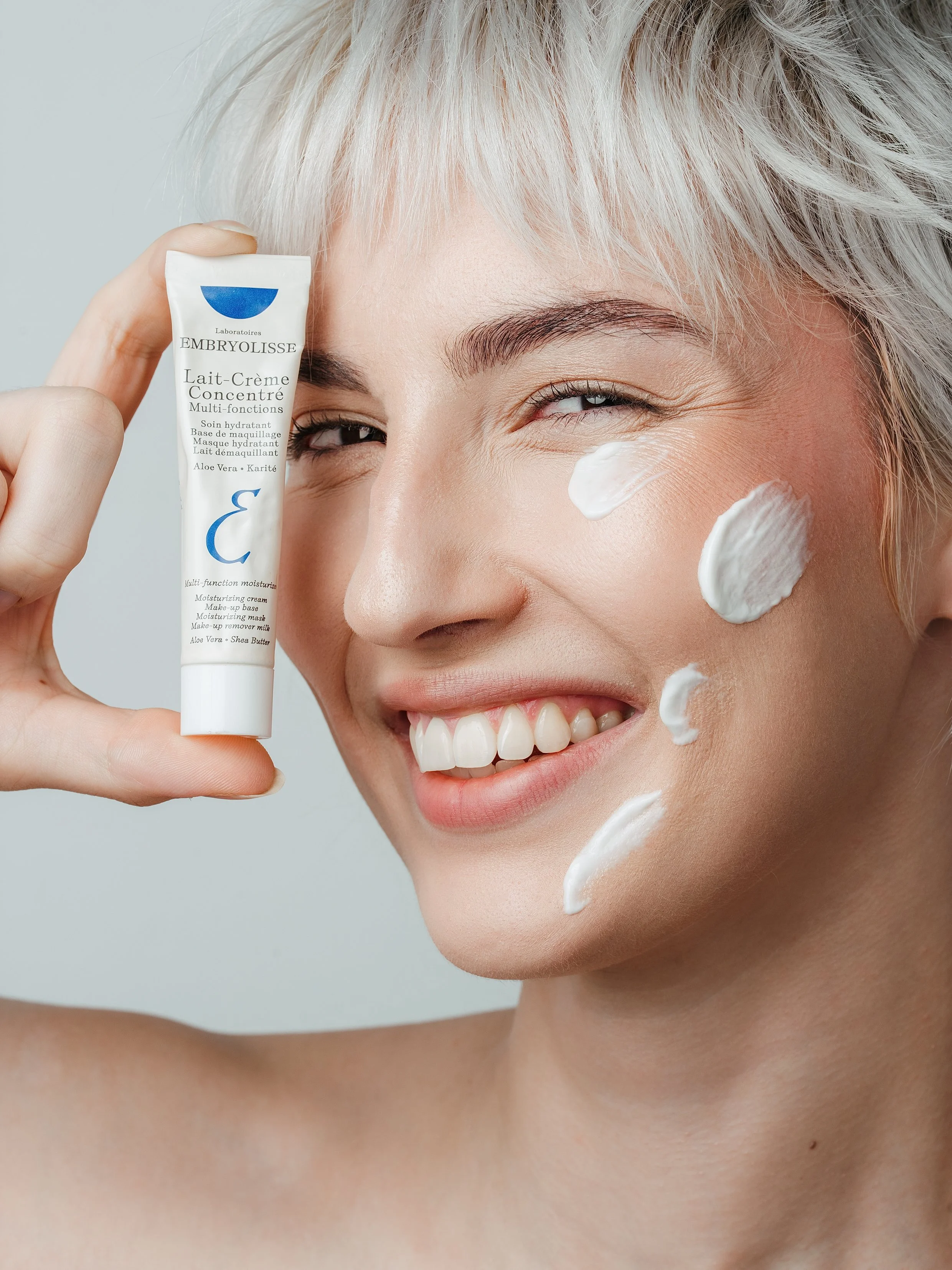 Une femme souriante avec des cheveux blonds courts, appliquant une crème hydratante sur son visage avec des spots de crème sur la joue. C'est un shooting beauté.