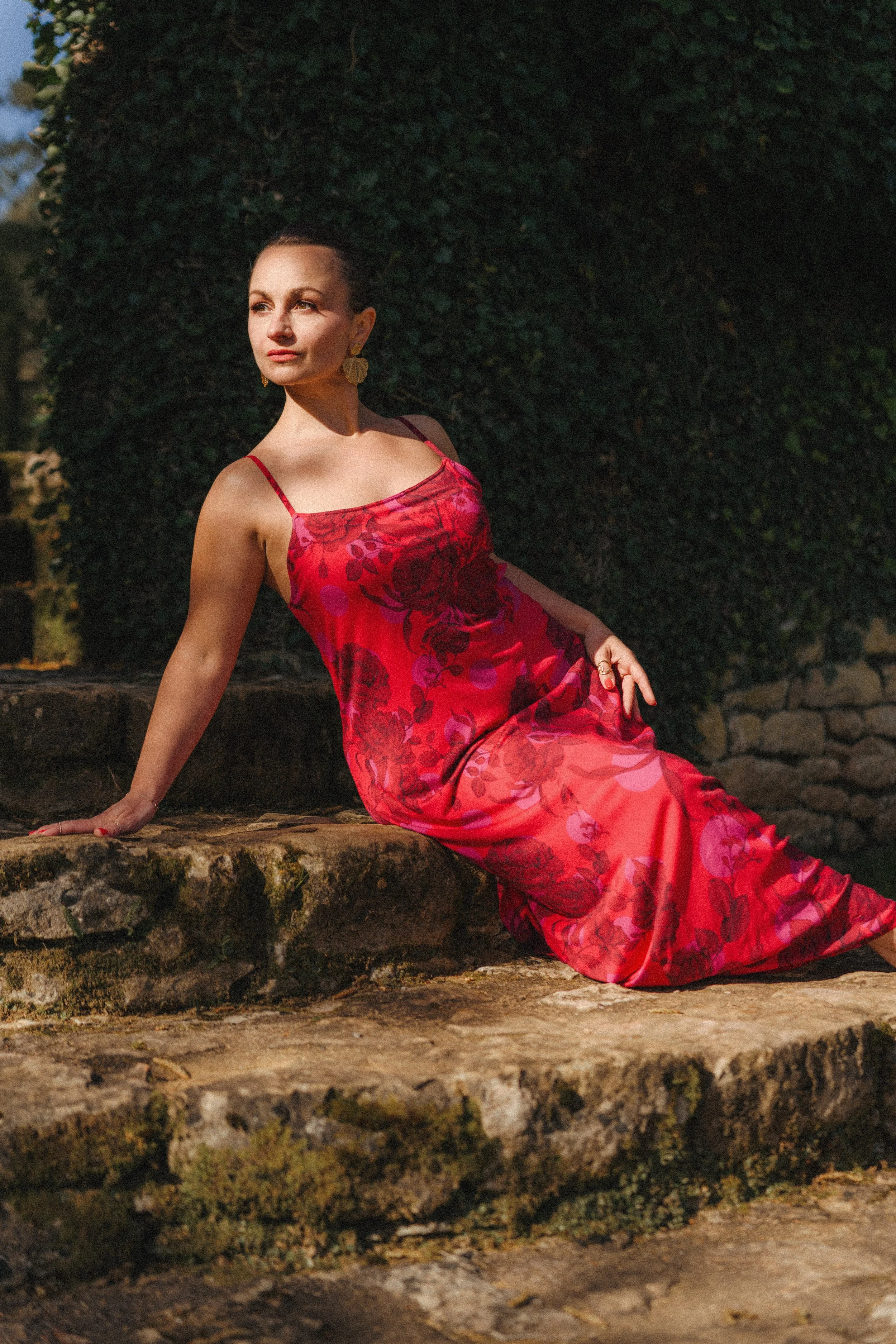 Femme portant une robe rouge avec des motifs floraux, assise sur des pierres, dans un jardin sous la lumière du soleil.