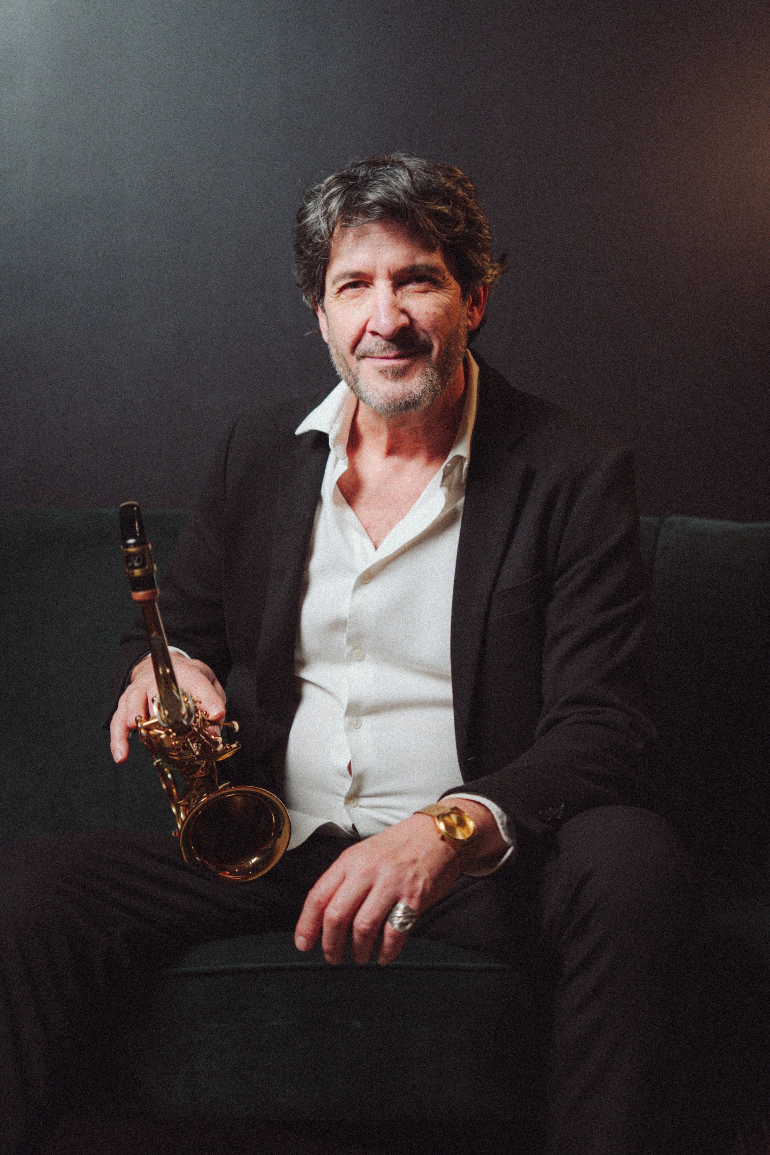 Un homme avec des cheveux grisonnants et une barbe, portant un costume noir, assis avec un saxophone dans la main, sur un fond sombre. Il porte une montre en or et une bague à la main gauche.