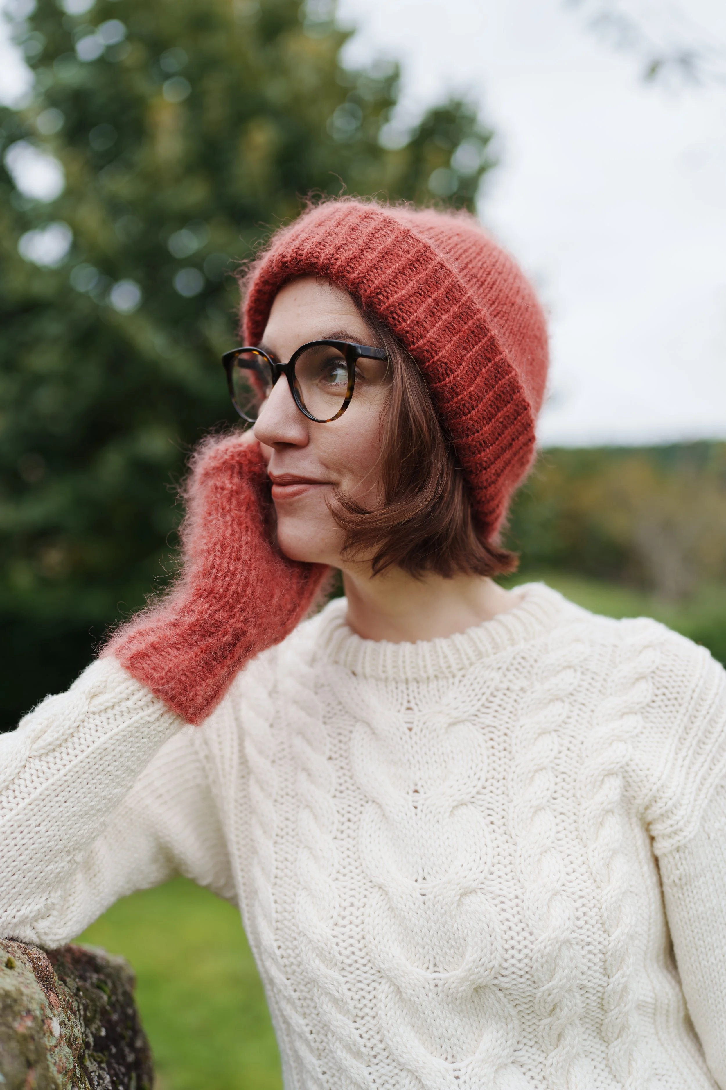 Une femme portant un bonnet en laine rouge, des lunettes écaille et une écharpe rose, avec un pull en maille blanc, dans un environnement extérieur avec des arbres en arrière-plan.
