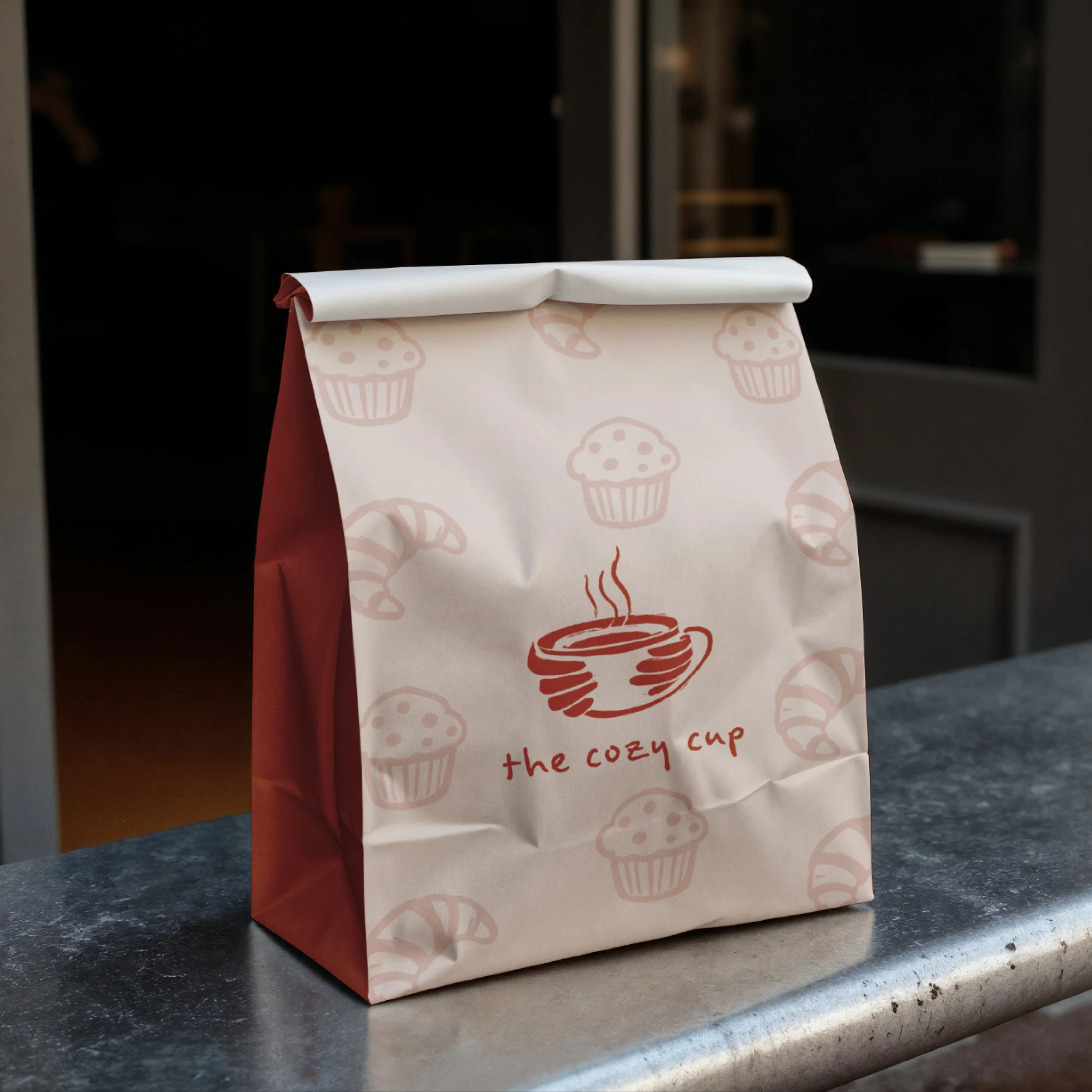 Eine designte To-Go Restaurant Tüte. Die Tüte ist bedruckt mit einem dampfenden Kaffeebecher und dem Text 'the cozy cup'. Das Design-Muster auf der Tüte besteht aus Cupcakes und Croissants.