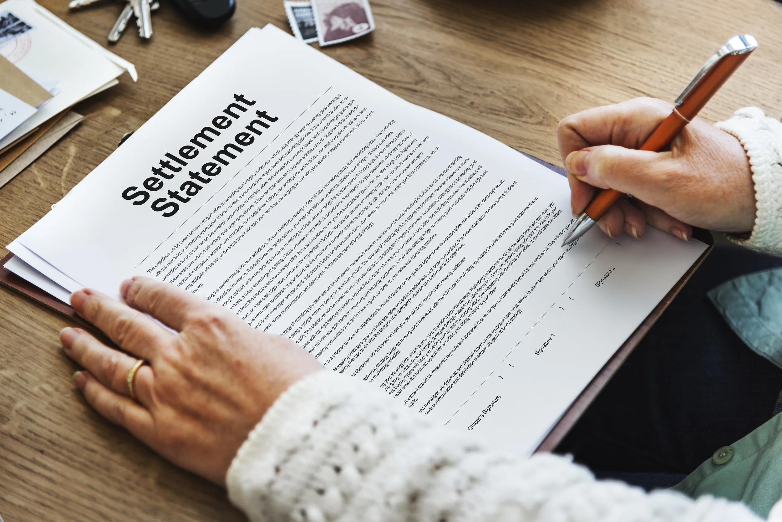 A person filling out a settlement statement document on a wooden desk, with a pen in hand, wearing a white sweater and a wedding ring.