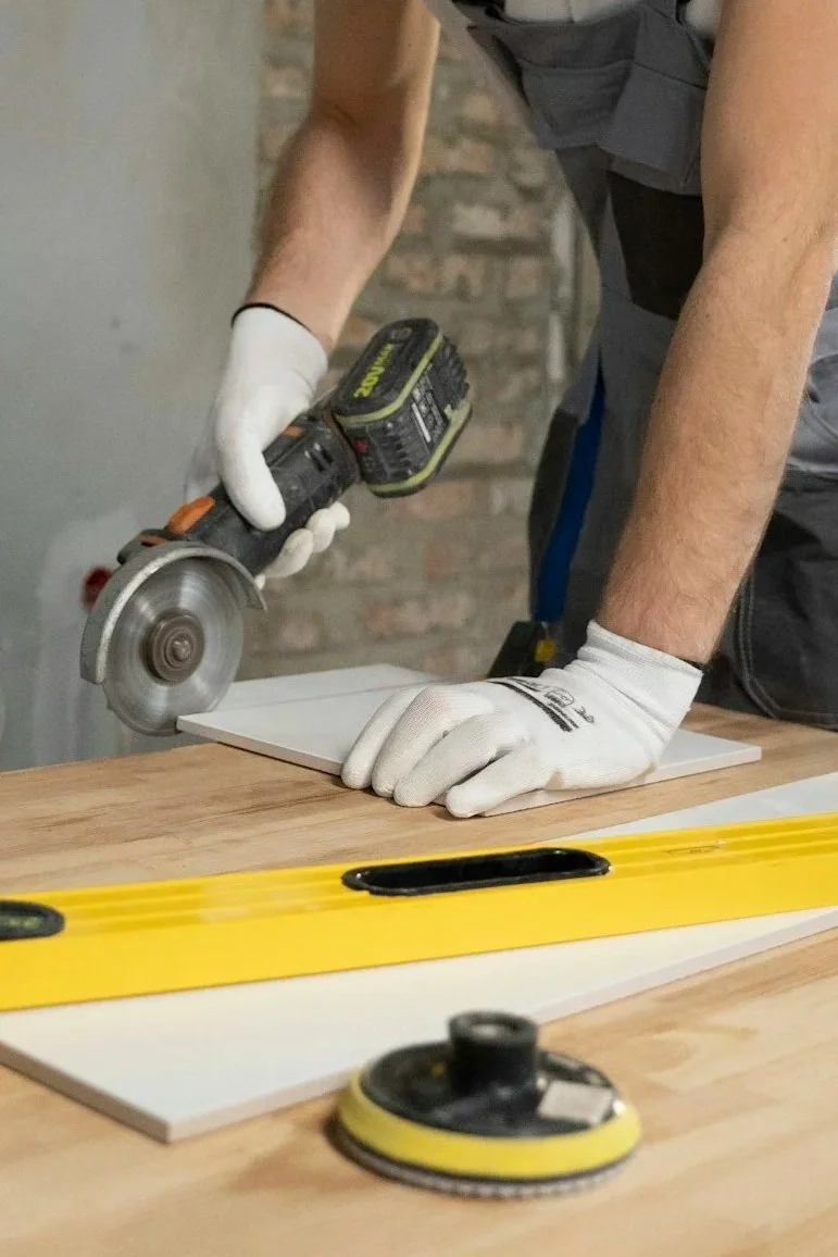 A person using a cordless circular saw to cut a piece of white wood on a workbench. The person is wearing white gloves, and there is a yellow level and a yellow and black sander or polisher on the work surface. The background shows a brick wall.