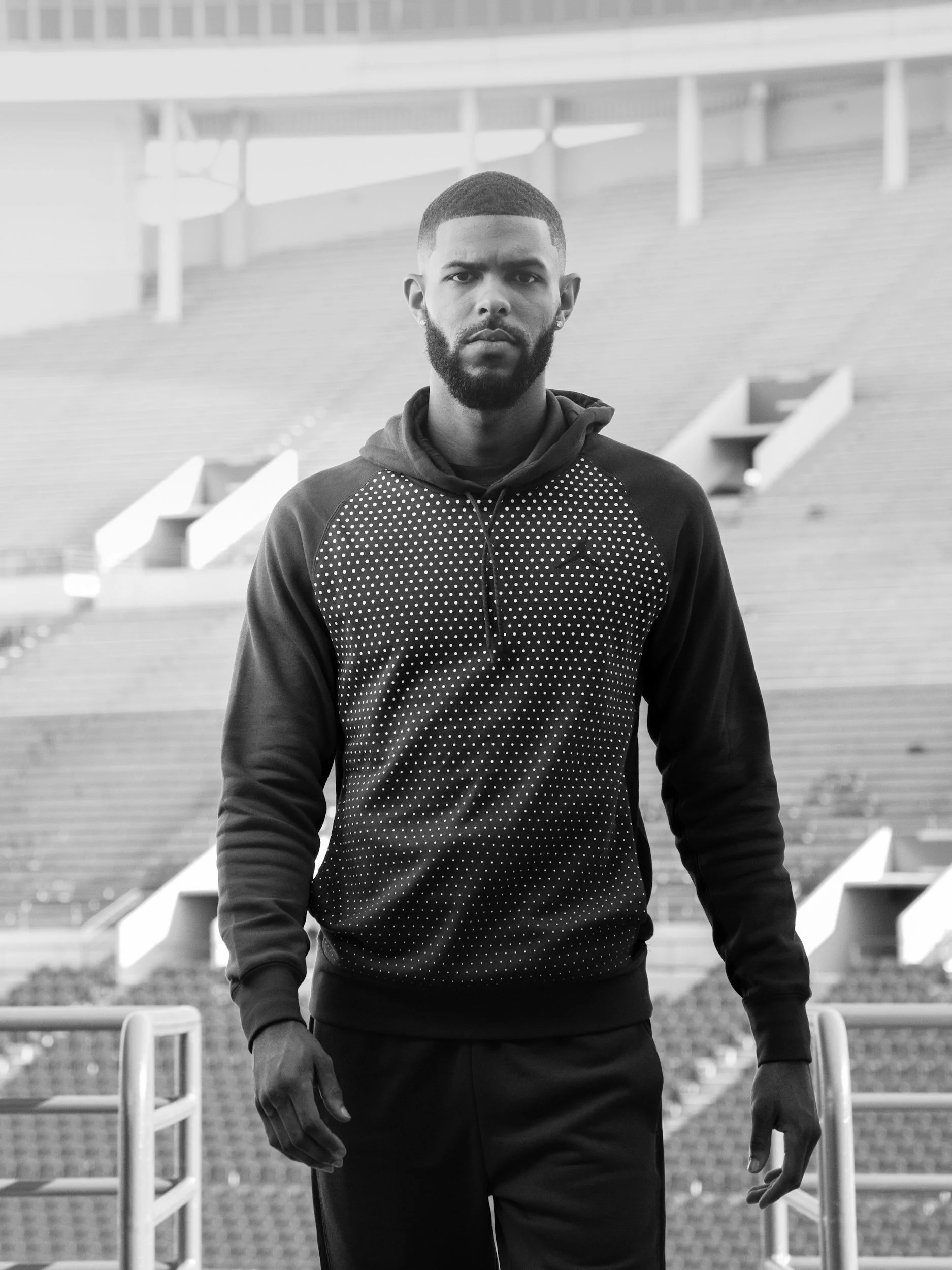 A man with a beard and short hair standing on stadium bleachers, wearing a dark hooded sweatshirt with a dotted pattern on the chest.