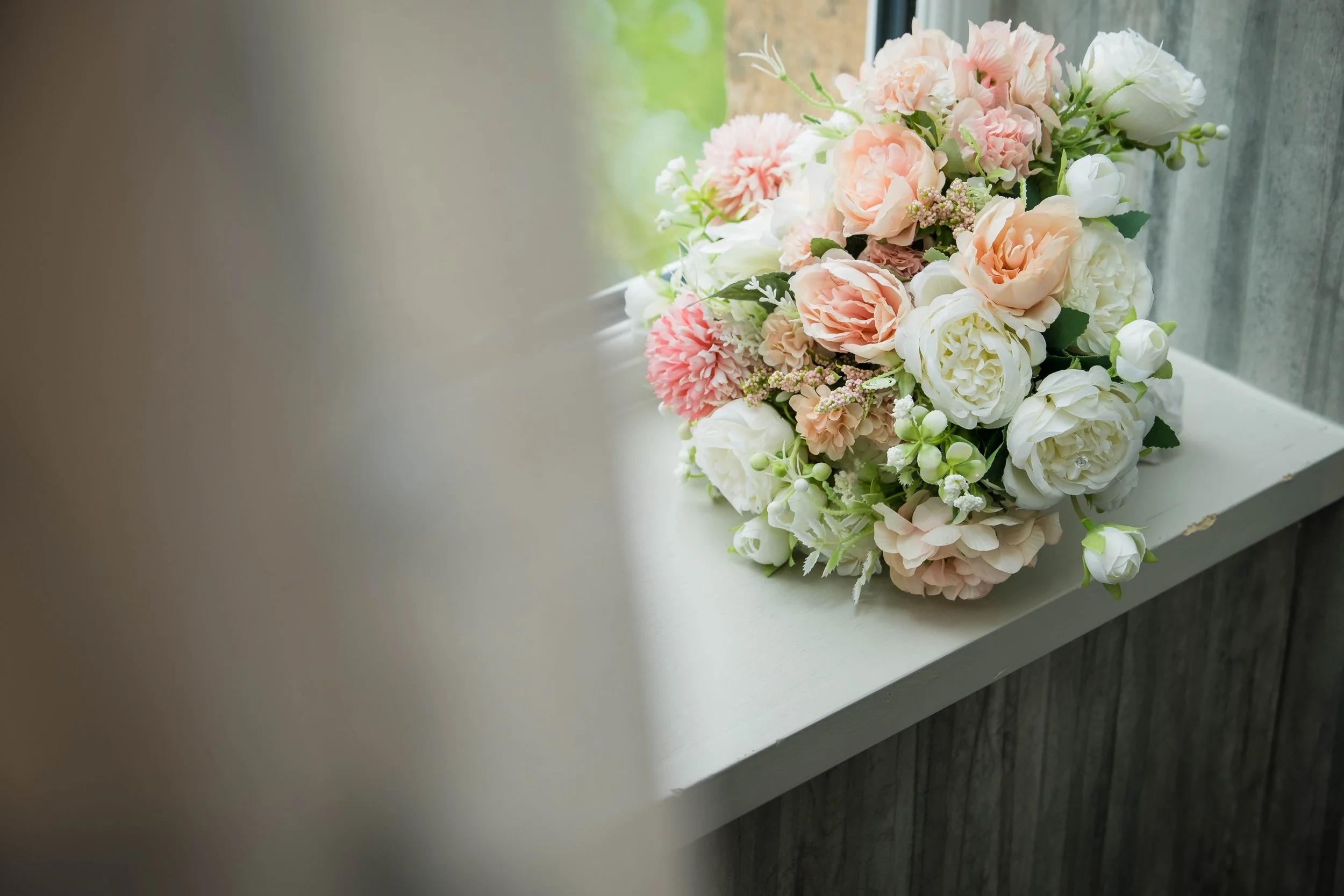 A bouquet of pink and white flowers resting on a white surface near a window with green foliage outside.