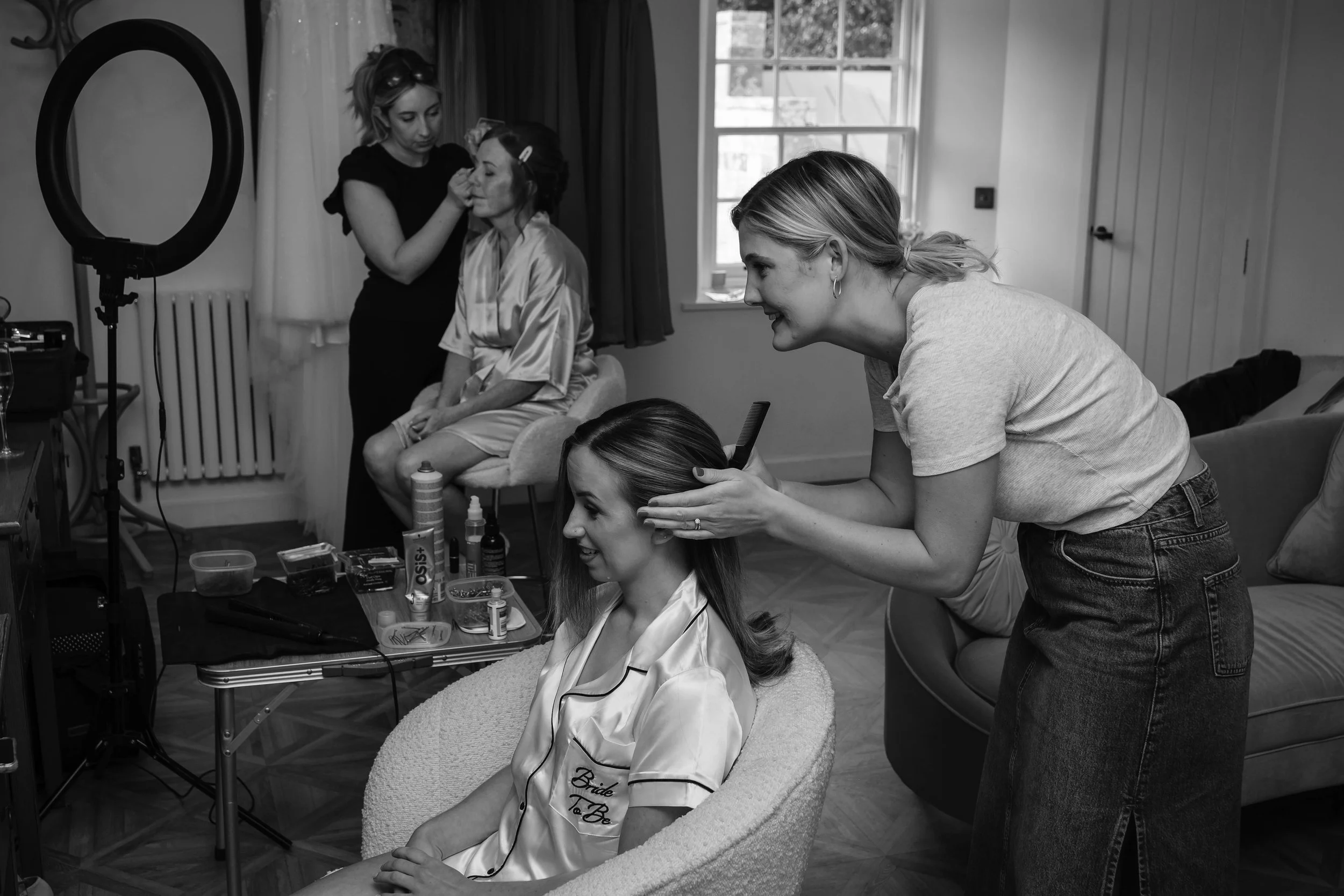 A bride sitting in a chair getting her hair styled by a professional while makeup artists work on another bride in the background, with a window and a ring light nearby.