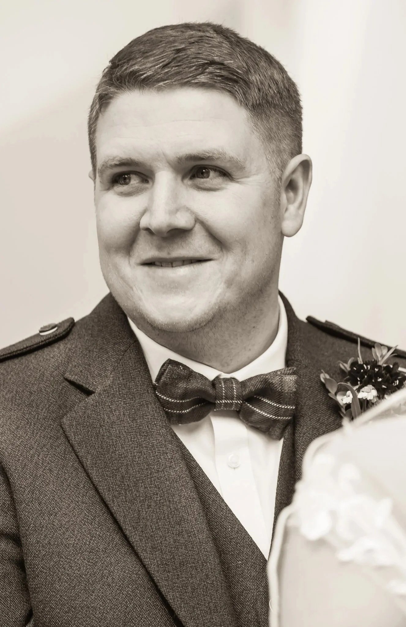 Black and white photo of a man in a suit, wearing a bow tie, smiling, with flowers partially visible on the right.