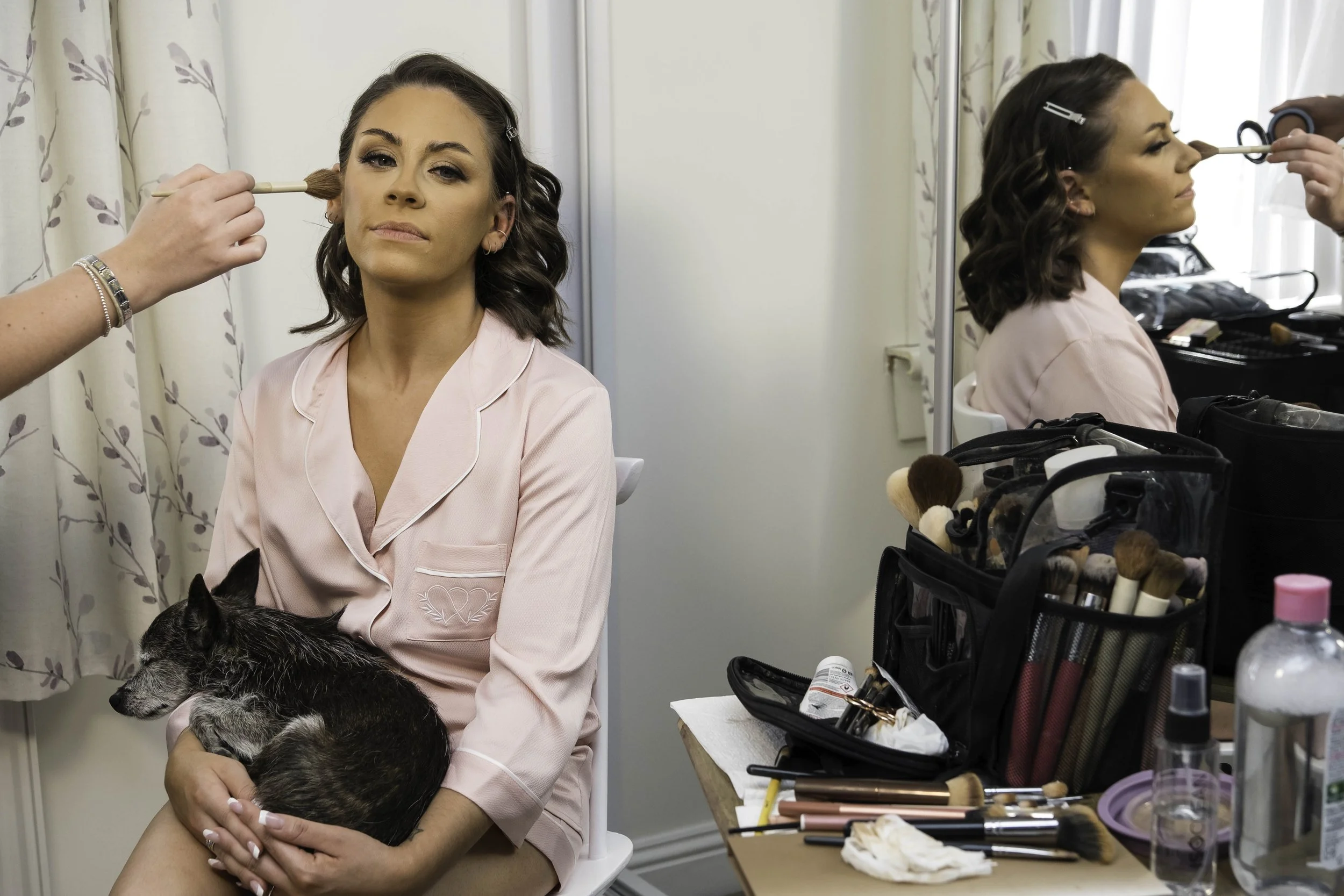 Woman in pink pajamas getting her makeup done, holding a dog, with makeup tools on a table nearby.