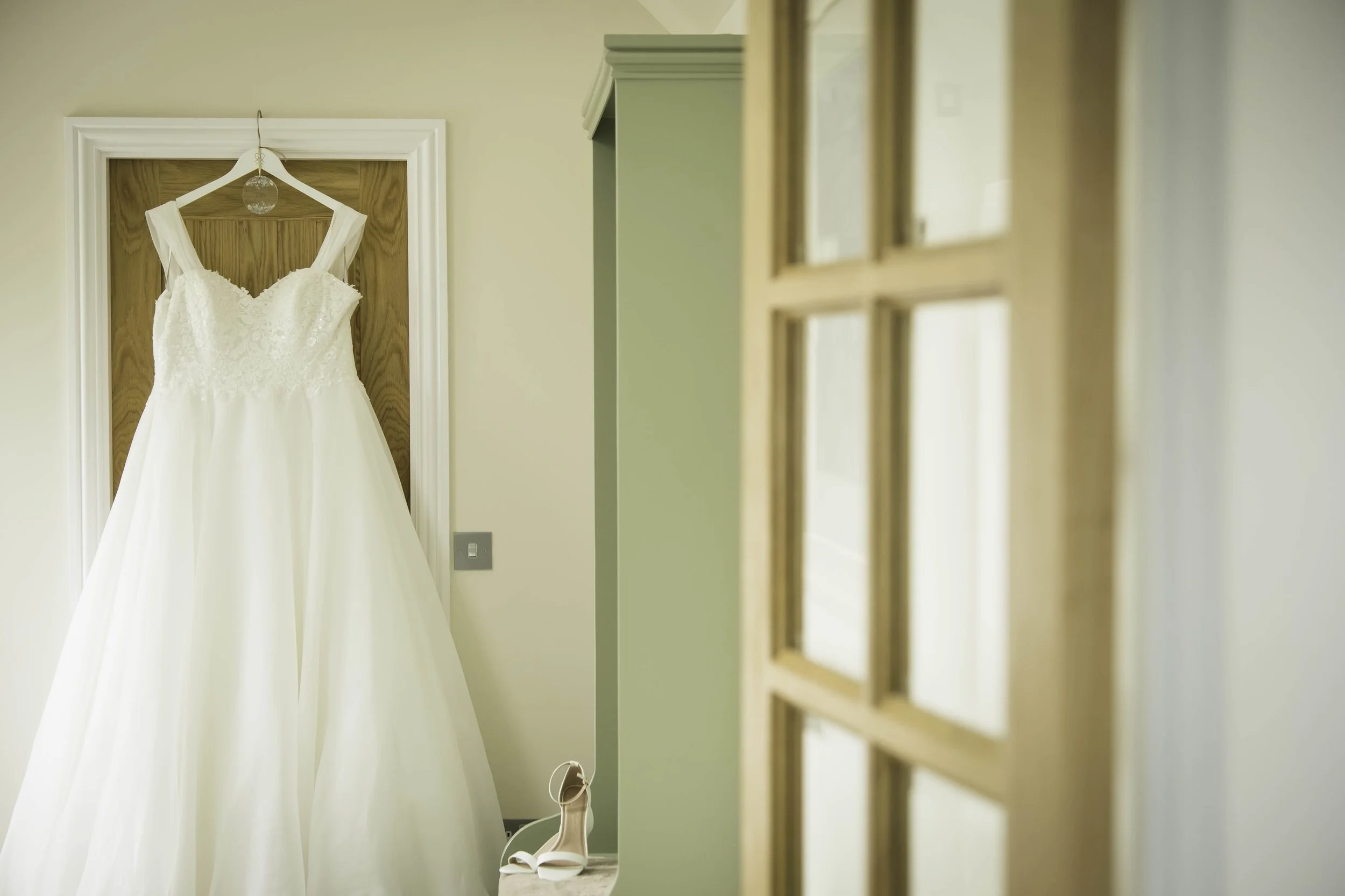A wedding dress hanging on a wooden door with a pair of white heels on a small table nearby.