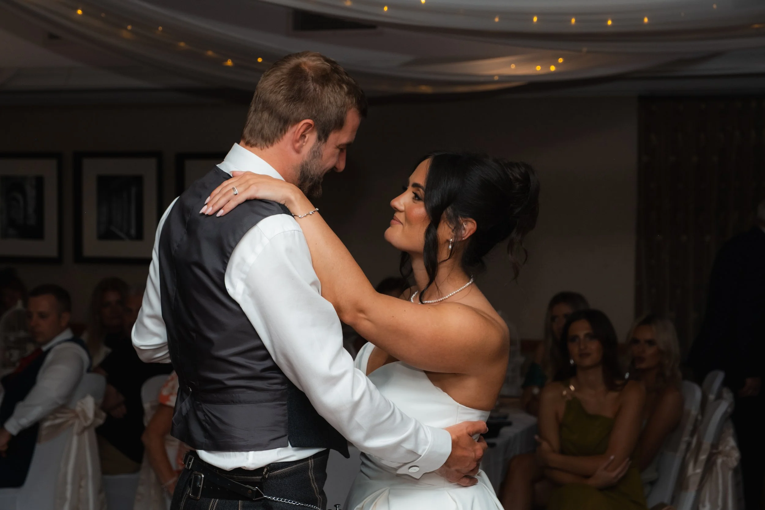 A newlywed couple sharing a dance at their wedding reception, surrounded by seated guests watching.