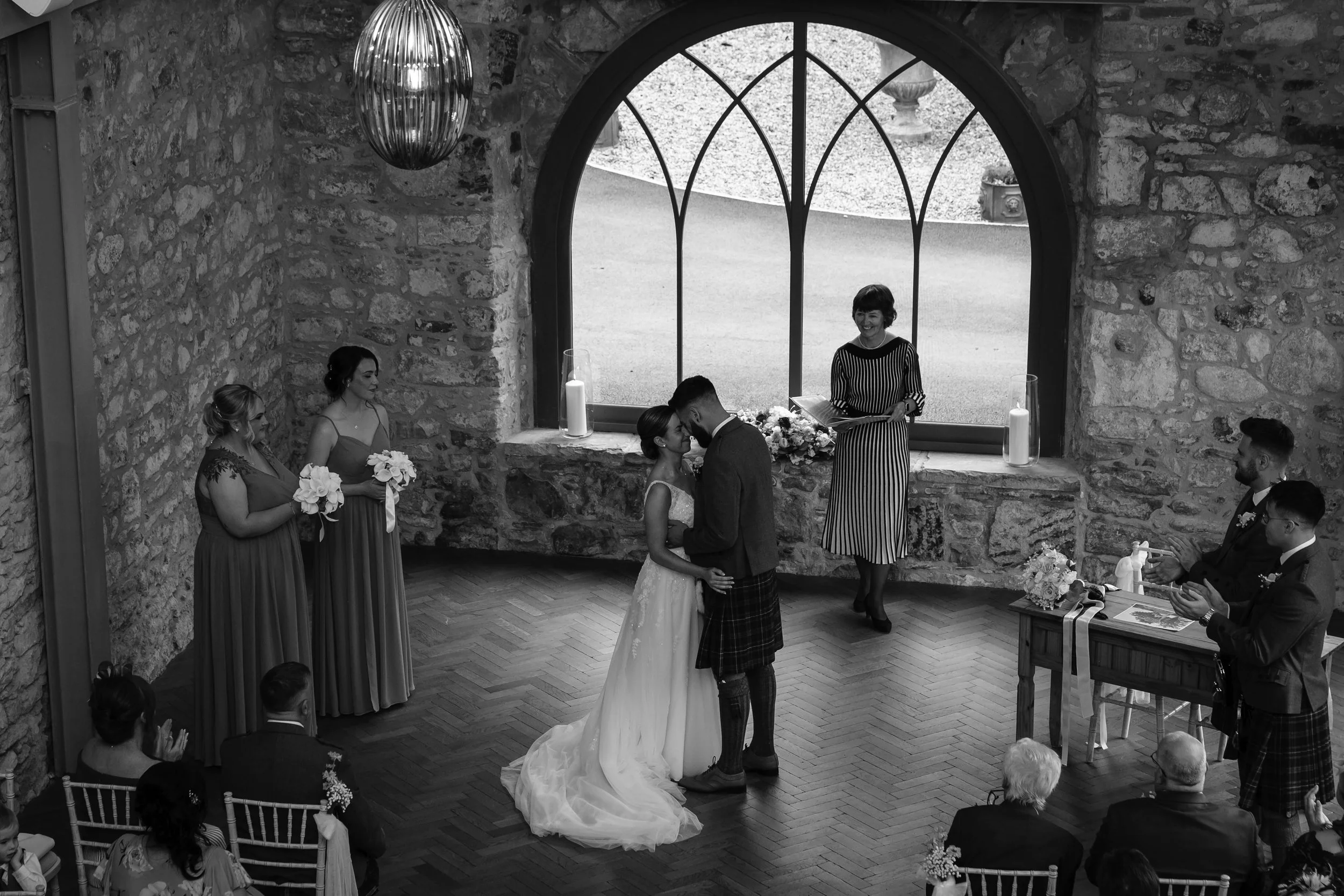 A wedding ceremony with a bride and groom kissing under a large window, surrounded by bridesmaids and groomsmen, with guests seated and clapping.
