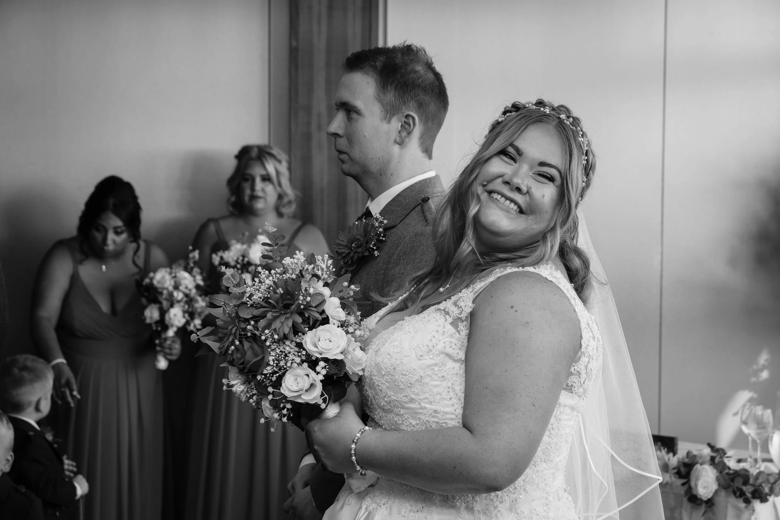Smiling bride in wedding dress holding bouquet with groom and bridesmaids in background.