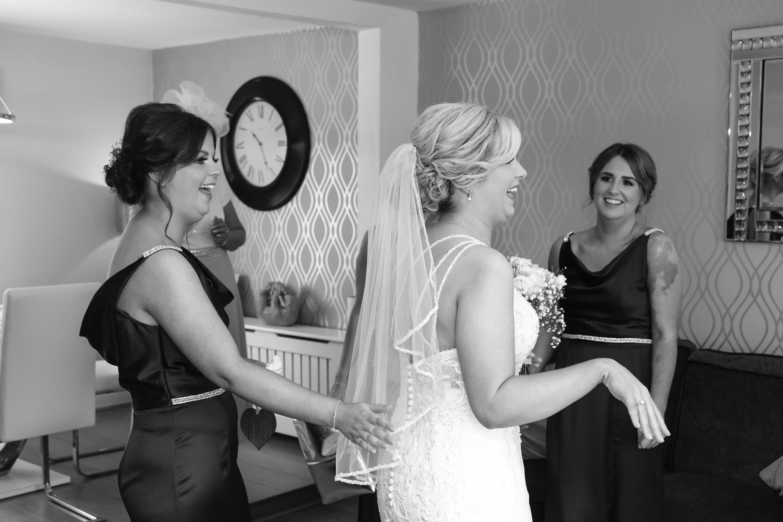 A bride in a wedding dress and veil laughing with three women in a living room during a wedding preparation.