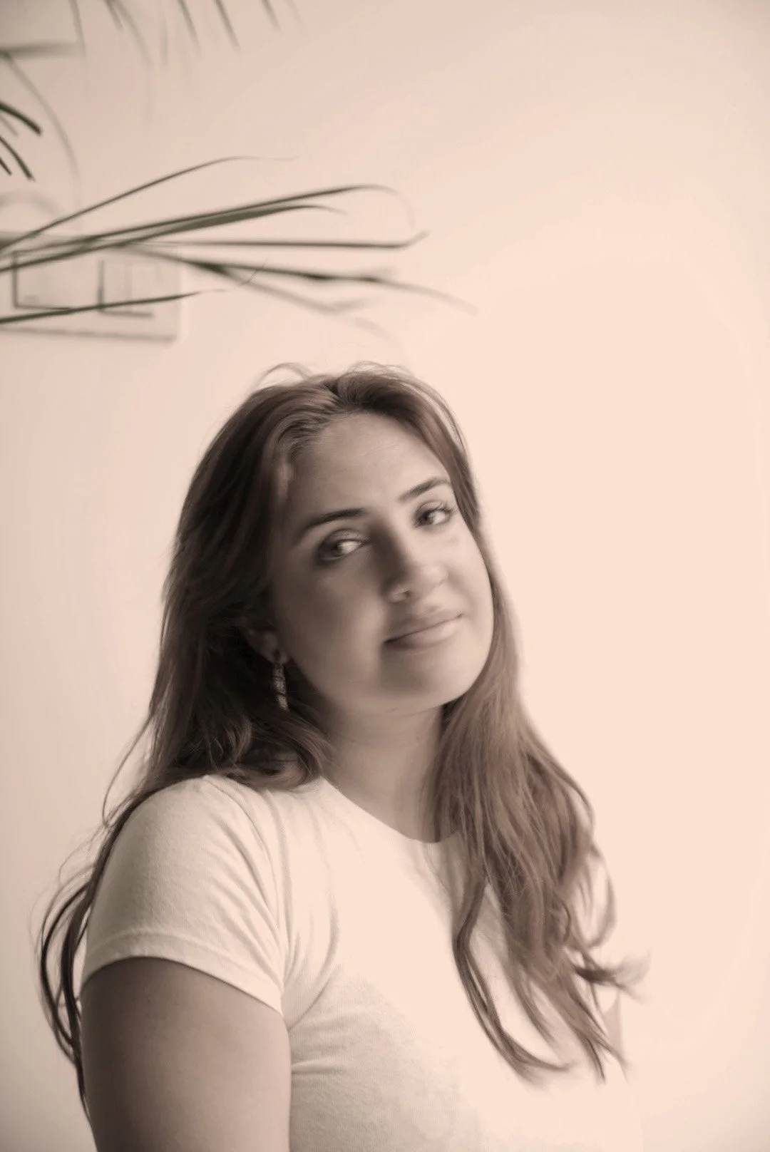 A young woman with long hair wearing a light-colored t-shirt, standing indoors against a plain wall, looking slightly to the side with a subtle smile.