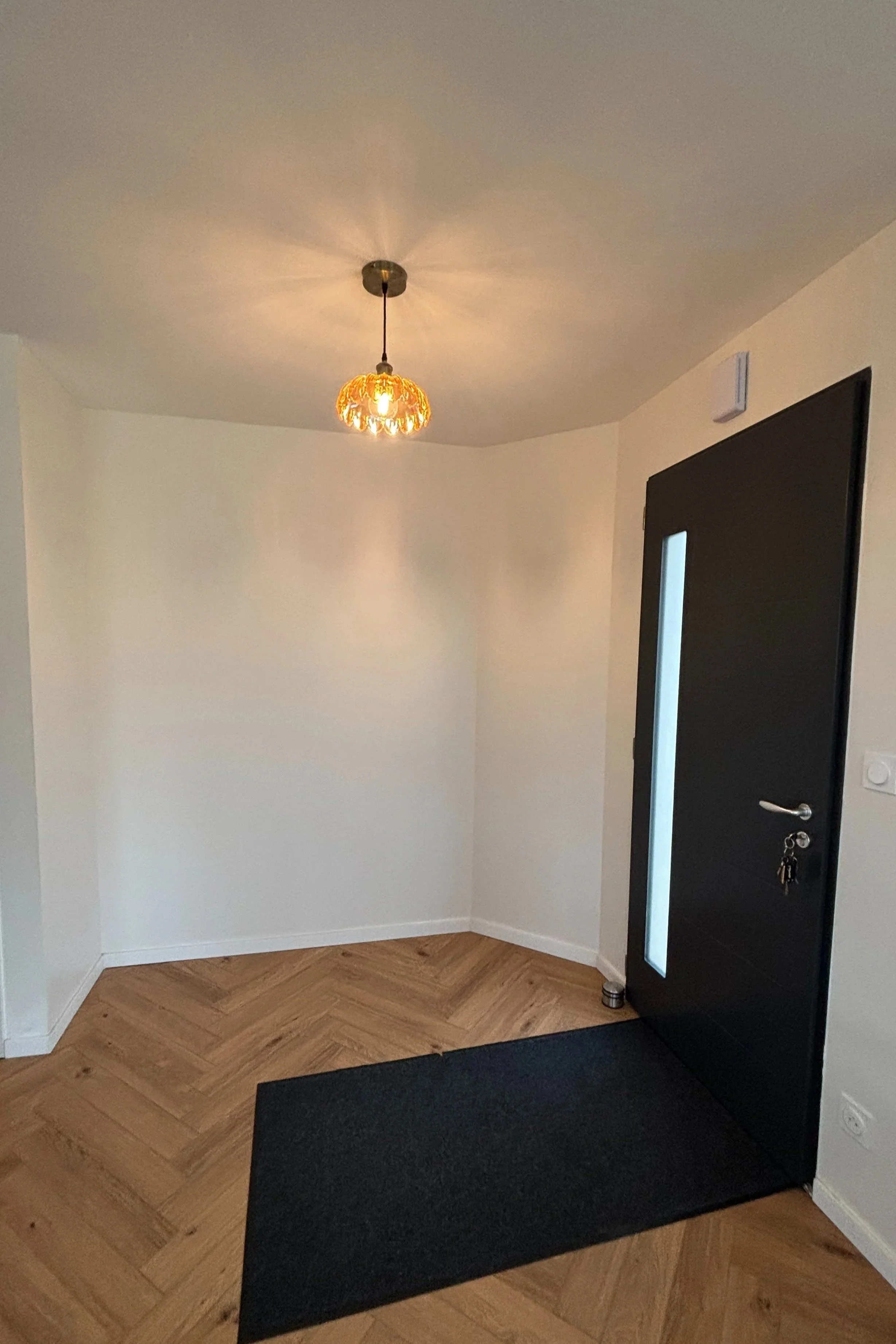 Vue de l'entrée d'une maison avec un sol en parquet en chevrons, une porte noire avec une vitre verticale, un tapis noir, un mur blanc, et un plafonnier suspendu avec un abat-jour en verre orange