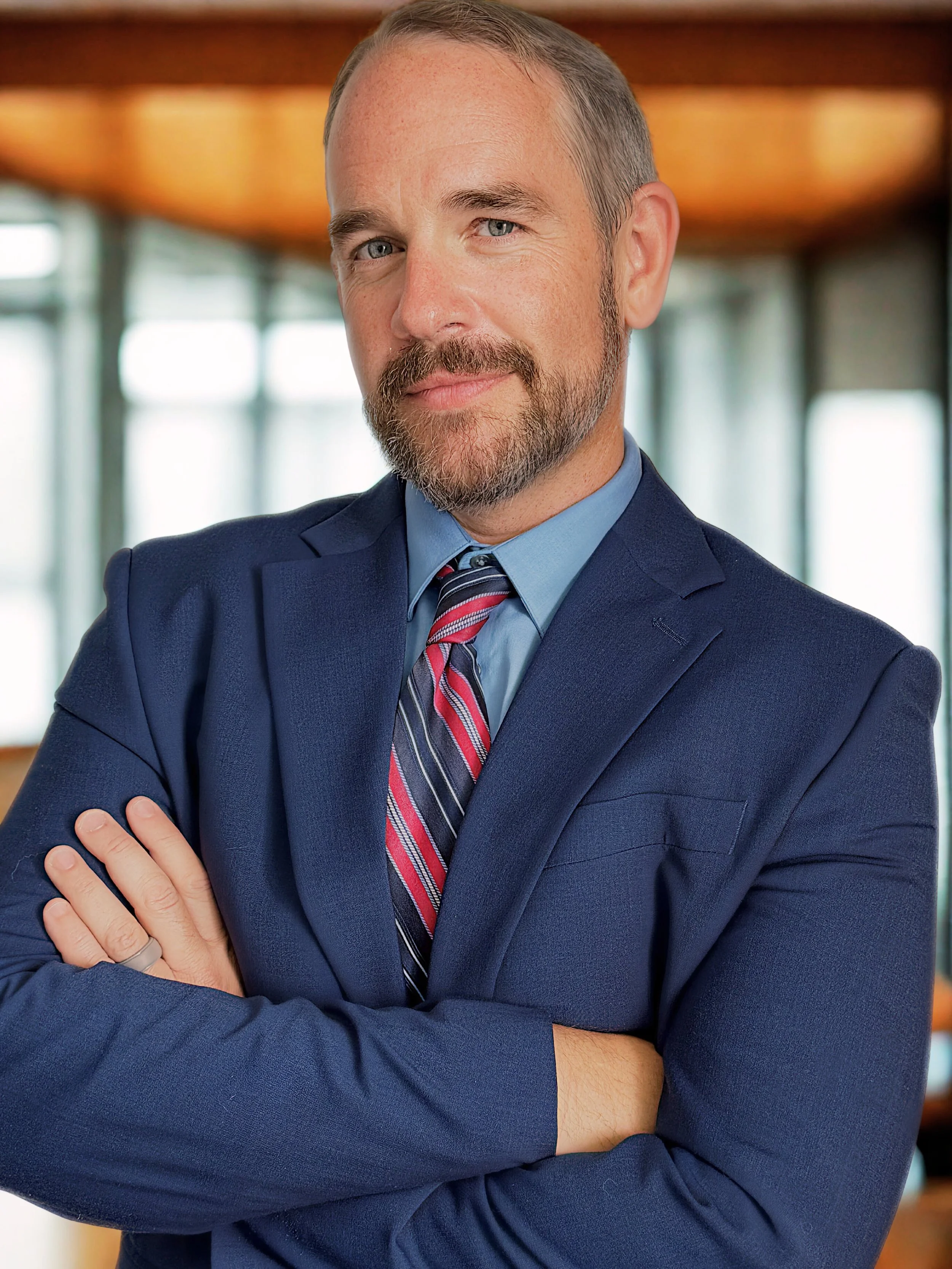 A professional man with a beard and light hair, wearing a navy suit, blue shirt, and striped tie, standing with arms crossed in an office setting.