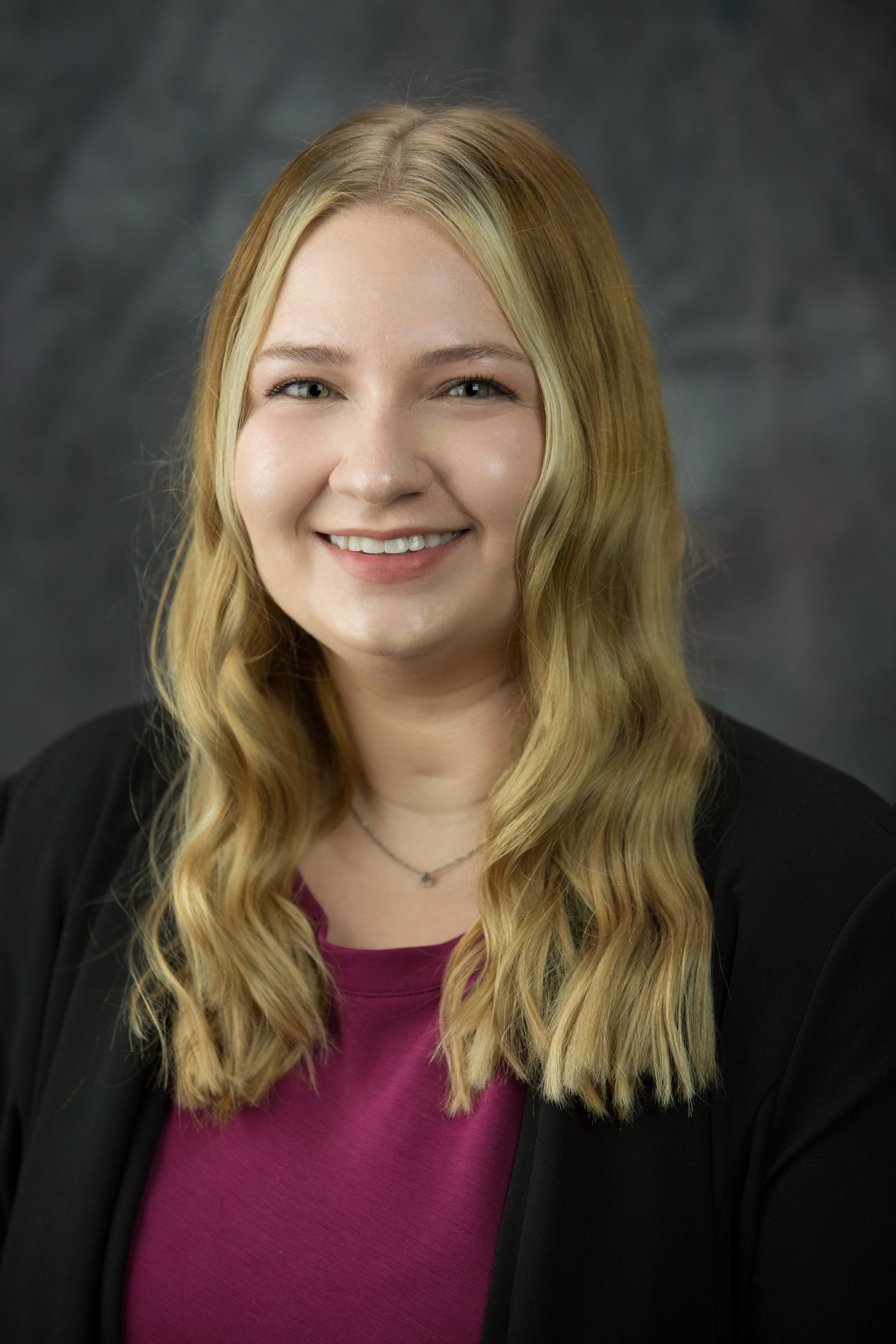A young woman with long, wavy blonde hair and blue eyes, smiling, wearing a black blazer over a maroon top, against a dark, blurry background.