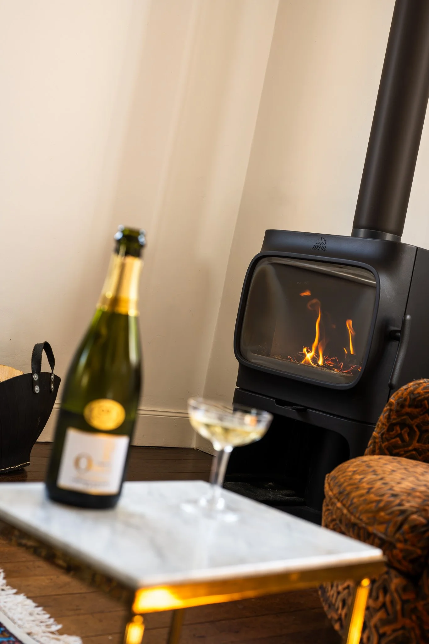 Une bouteille de champagne, deux verres à vin remplis, un poêle à bois avec des flammes, un fauteuil en tissu imprimé, un tapis blanc, une table en marbre, un mur beige.